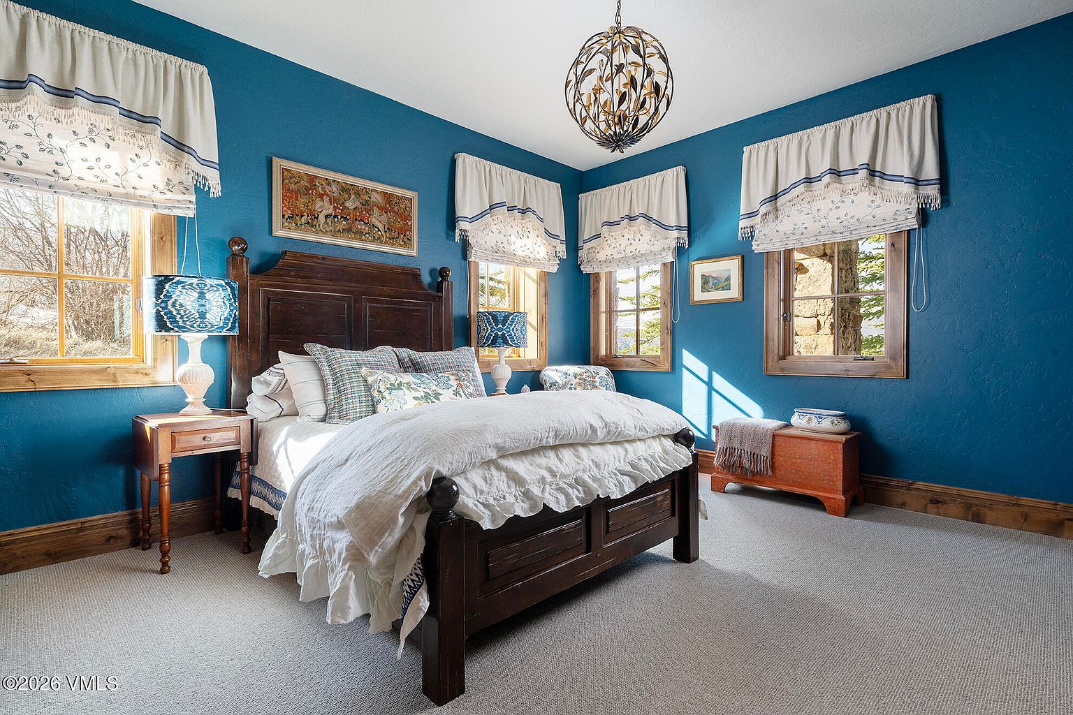 This cozy bedroom features bold blue walls contrasted by a dark wood bed frame and matching side table. The room is illuminated by a unique, spherical metal chandelier and natural light streaming through multiple windows dressed in patterned valances. A wooden chest sits at the foot of the bed, adding a rustic touch to the space, which is finished with neutral carpeting.