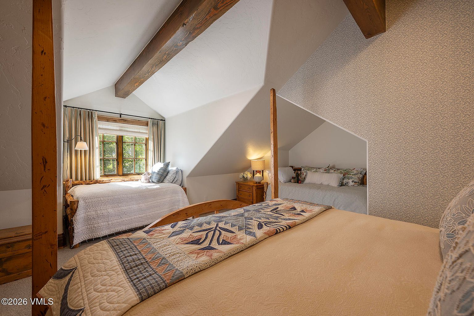 This cozy, rustic-style bedroom features a multi-bed layout with a primary bed in the foreground and two additional twin beds tucked into alcoves. The room is characterized by exposed wooden ceiling beams, sloped walls, and a warm, inviting atmosphere with patterned quilts and soft textiles. The perspective is from the foot of the main bed, looking toward the windowed alcove and the secondary sleeping nook.