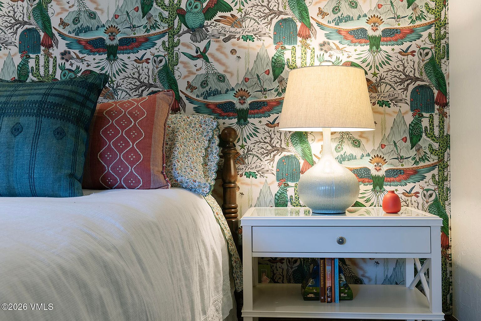 This cozy bedroom corner features a bed dressed in a white textured quilt and a mix of patterned throw pillows, set against a vibrant, whimsical wallpaper depicting desert landscapes and wildlife. A white nightstand with a glass top holds a ceramic table lamp with a soft, warm glow and a small orange decorative object. The scene is captured from a close-up, eye-level perspective, highlighting the eclectic and playful design aesthetic of the room.