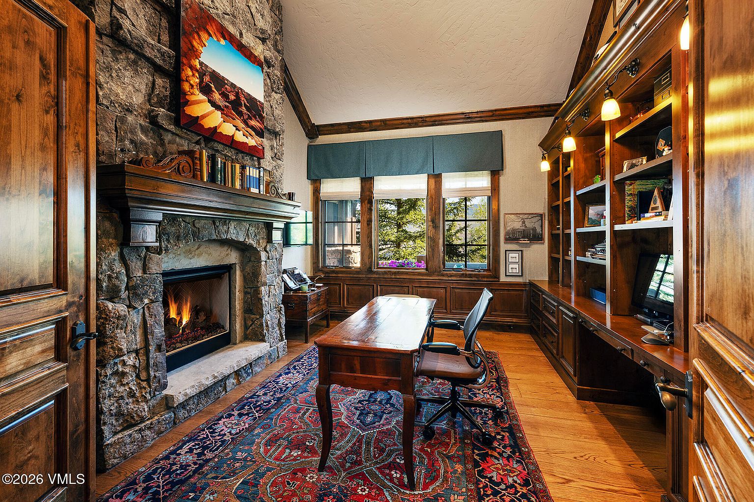 This sophisticated home office features a prominent stone-clad chimney breast with a built-in fireplace and a decorative mantel. The room is anchored by a large wooden desk and a patterned area rug, complemented by extensive custom wood shelving and cabinetry along the wall. Large windows with blue valances provide natural light and views of the outdoors, creating a warm and professional atmosphere.