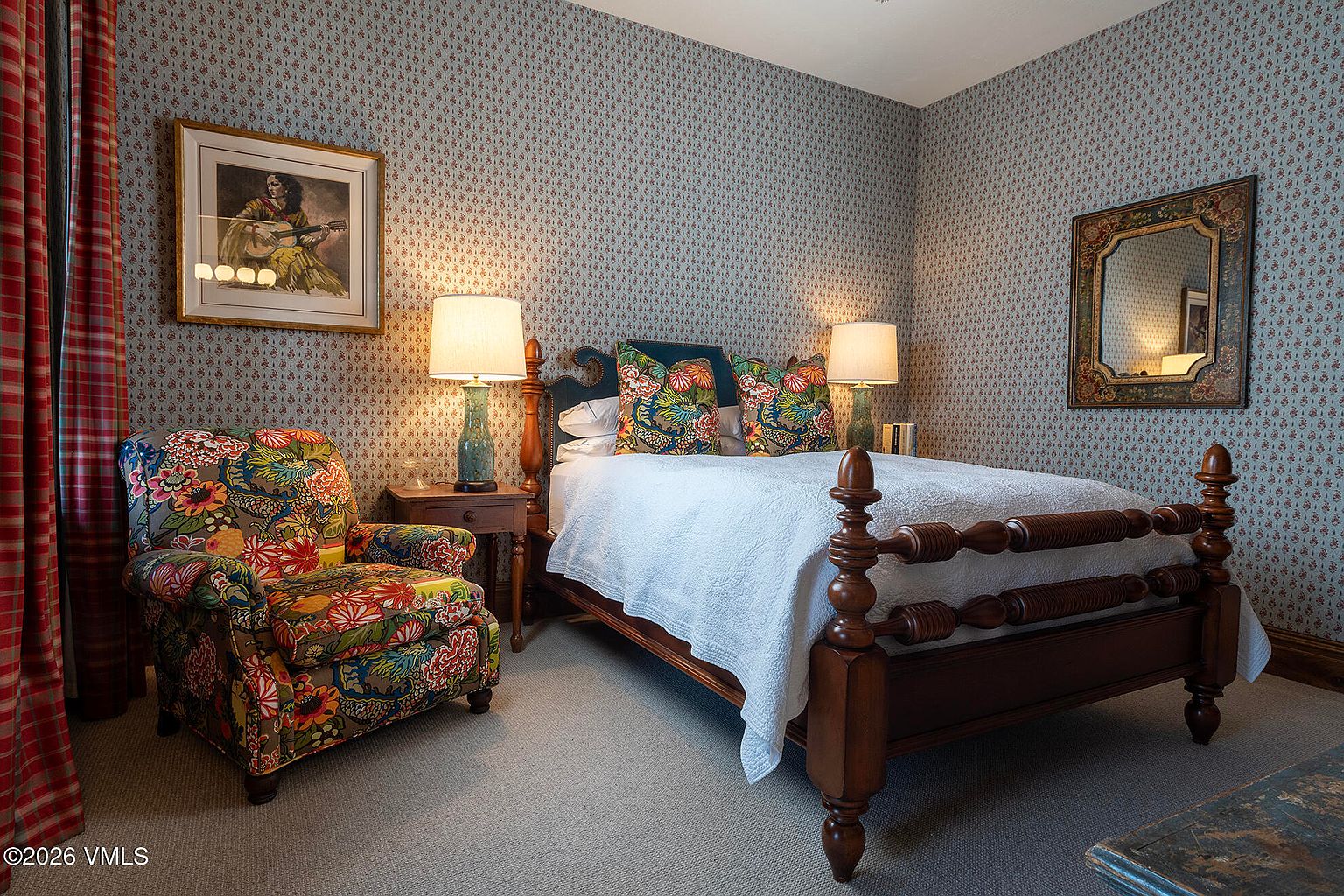 This cozy guest bedroom features a traditional four-poster wooden bed dressed in a white quilt and vibrant floral-patterned accent pillows. The room is decorated with patterned wallpaper, a matching floral armchair, and a framed piece of art, creating a warm and classic aesthetic. A wooden nightstand with a lamp sits beside the bed, while a decorative mirror hangs on the adjacent wall, adding to the room's inviting and well-appointed atmosphere.