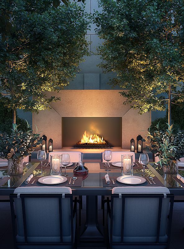 This elegant outdoor dining space features a formal table set for four, positioned directly in front of a large, stone-clad fireplace. The scene is framed by two lush, illuminated trees that create an intimate, symmetrical atmosphere. The composition is captured from a centered, eye-level perspective, highlighting the sophisticated lighting and refined furniture design.