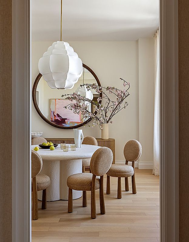This elegant dining room is captured from a doorway perspective, showcasing a sculptural, light-toned pedestal table surrounded by four rounded, upholstered chairs. A large, circular mirror reflects the space, positioned above a textured sideboard that holds a tall vase with flowering branches. A prominent, white, multi-faceted pendant light hangs above the table, creating a sophisticated and serene atmosphere with a neutral color palette.