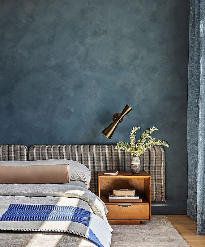 This sophisticated bedroom features a deep blue textured accent wall behind a plaid upholstered headboard. A warm-toned wooden nightstand sits beside the bed, topped with a decorative vase and a modern brass wall-mounted sconce. The bed is dressed in neutral linens with a vibrant blue and grey patterned throw, creating a cozy and stylish atmosphere.
