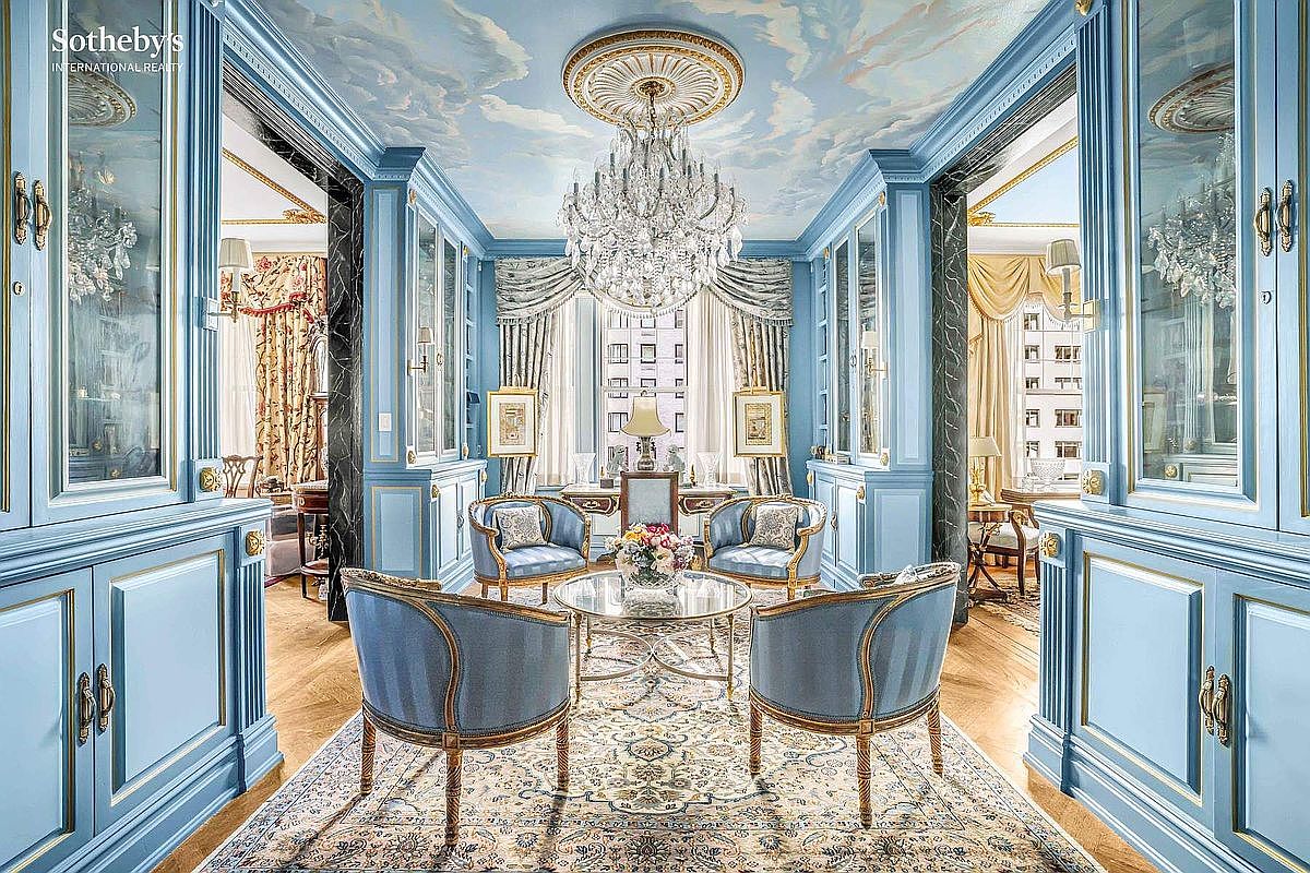 This is an elegant living room featuring light blue walls and cabinetry with gold trim, a cloud-painted ceiling with an ornate chandelier, and a patterned rug. Four blue upholstered chairs surround a glass coffee table, creating a sophisticated and inviting space. The room is flanked by mirrored cabinets, enhancing the sense of spaciousness and luxury.