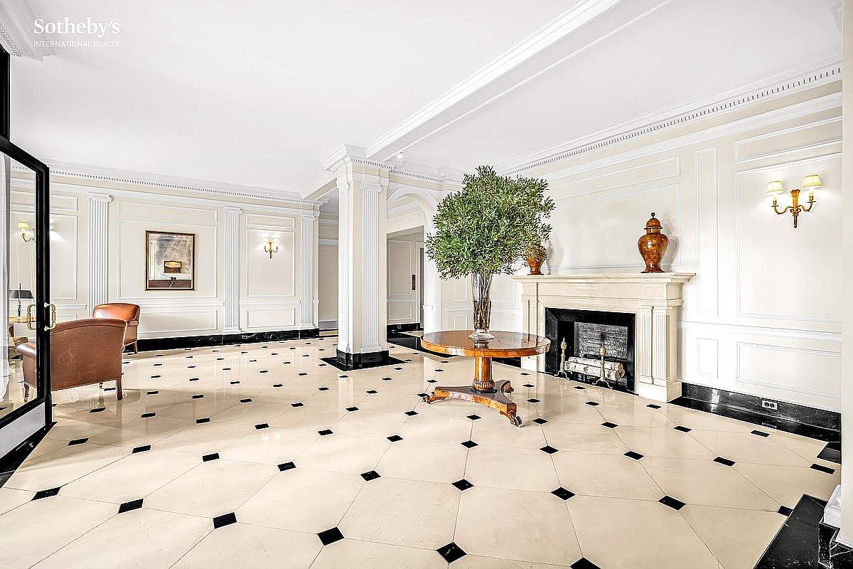This is an interior shot of a grand hallway featuring a classic design with white paneled walls, marble flooring with black accents, and elegant architectural columns. A round wooden table with a large plant sits in the center, and a fireplace adds a touch of warmth and sophistication. The perspective is from a wide angle, showcasing the spaciousness and luxurious feel of the entrance area.