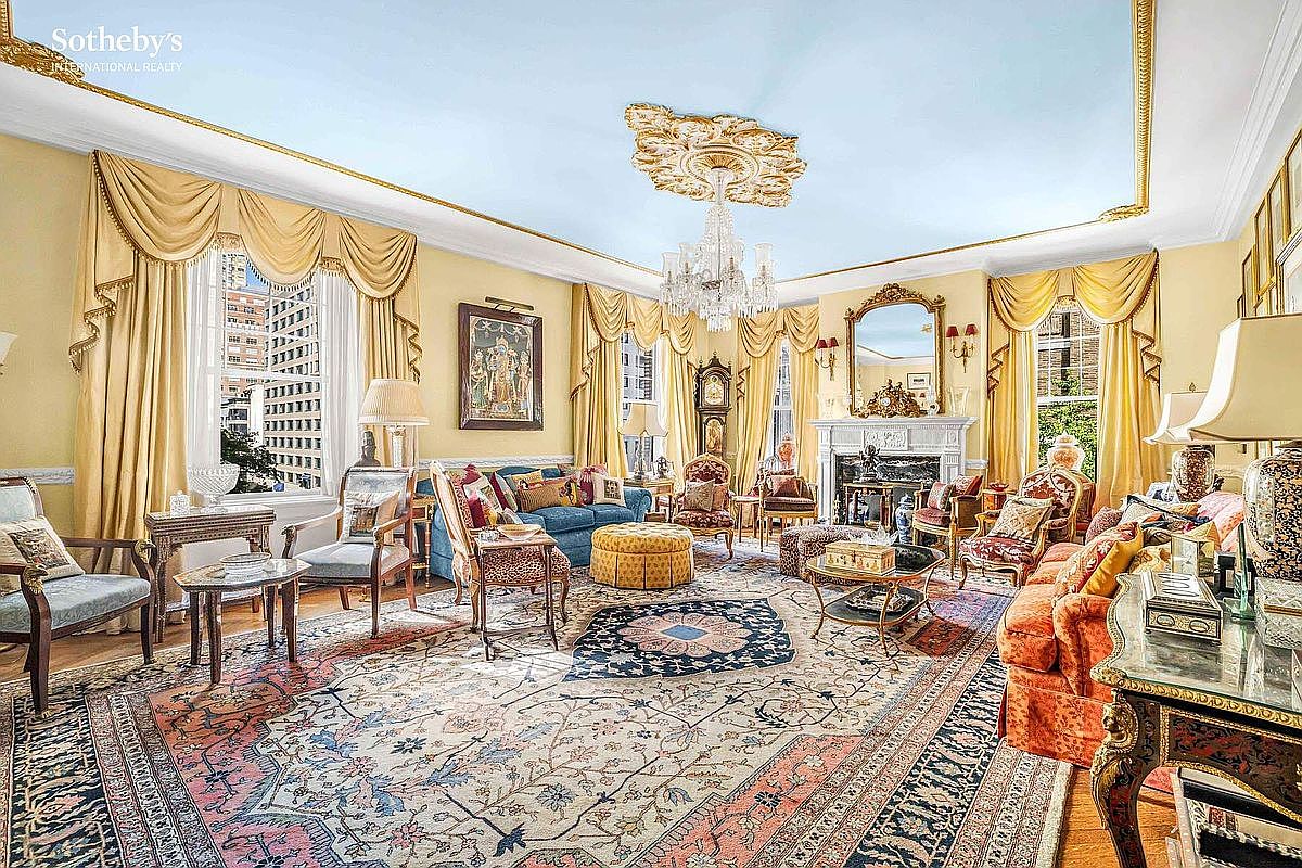 This is a luxurious living room featuring a mix of classic and ornate decor. The room is filled with antique furniture, including sofas, chairs, and tables, all arranged on a large, intricately patterned rug. A chandelier hangs from the ceiling, and the windows are draped with elegant curtains, creating a warm and inviting atmosphere.