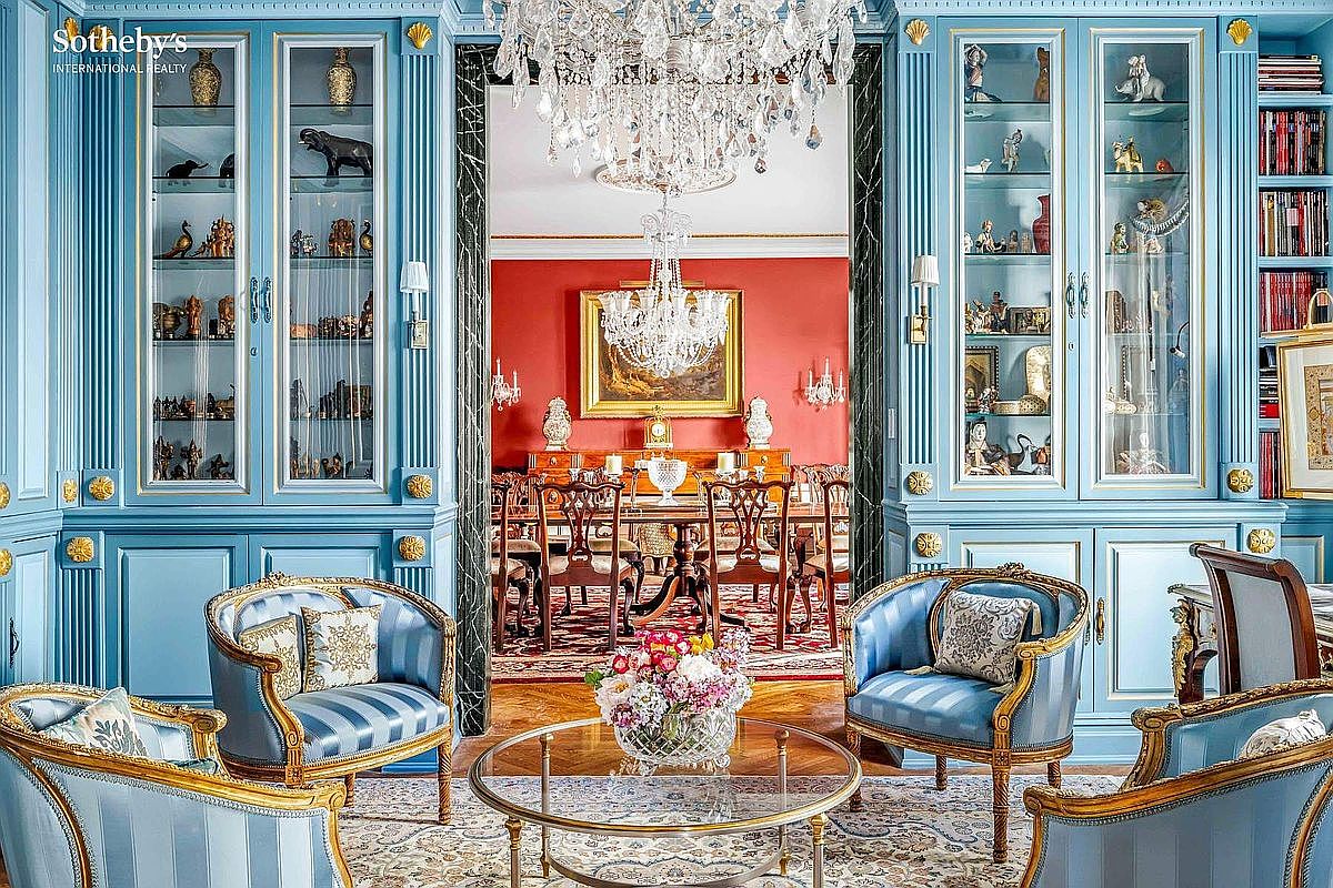 This opulent living room showcases a sophisticated design with custom blue cabinetry, elegant armchairs with striped upholstery, and a crystal chandelier. The room opens into a formal dining area with a red accent wall, creating a seamless flow for entertaining. A glass-topped coffee table sits atop an ornate rug, adding to the room's luxurious ambiance.