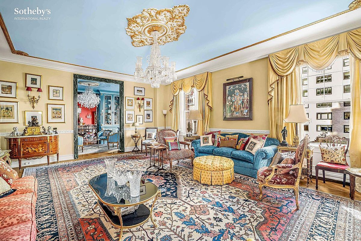 This is an opulent living room featuring a light blue ceiling, ornate gold trim, and a large crystal chandelier. The room is furnished with a blue velvet sofa, patterned chairs, and a detailed oriental rug. The space exudes luxury and classic elegance, perfect for showcasing a high-end property.