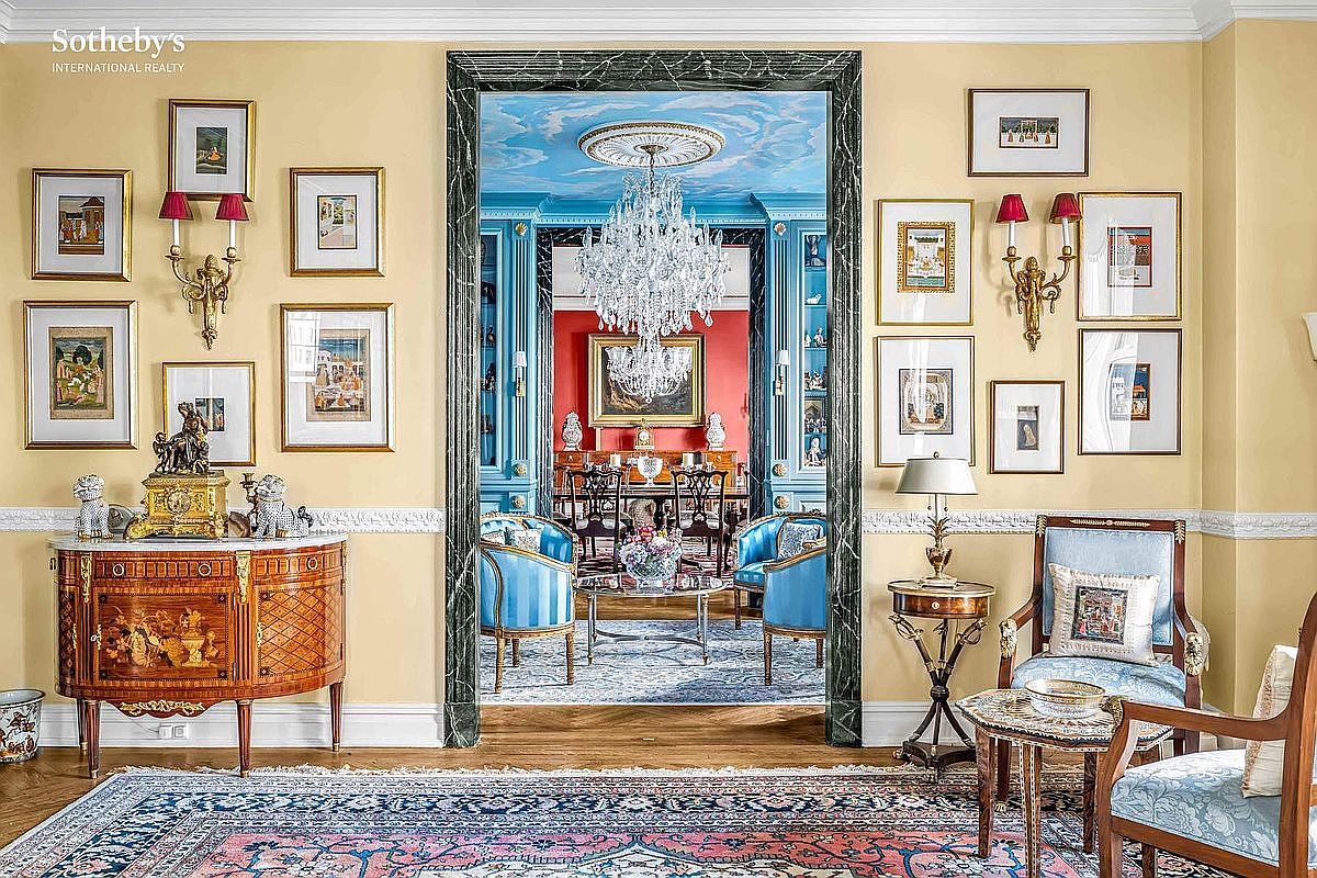 This interior shot showcases a luxurious living room that transitions into a dining area through a grand doorway. The living room features elegant furnishings, including blue upholstered chairs, a decorative rug, and walls adorned with framed artwork. The dining area beyond boasts a crystal chandelier and a formal dining set, creating an impression of sophisticated living and seamless flow between spaces.