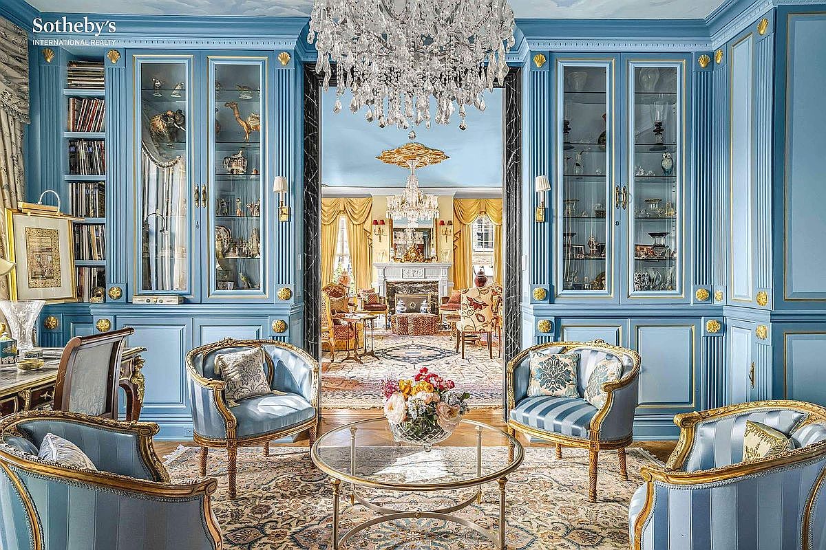 This is an elegant living room featuring light blue walls with gold accents, built-in display cabinets, and a crystal chandelier. The room is furnished with ornate chairs and a glass coffee table, all arranged on a patterned rug. The view extends through an open doorway to another elegantly decorated room with a fireplace, creating a sense of spaciousness and luxury.