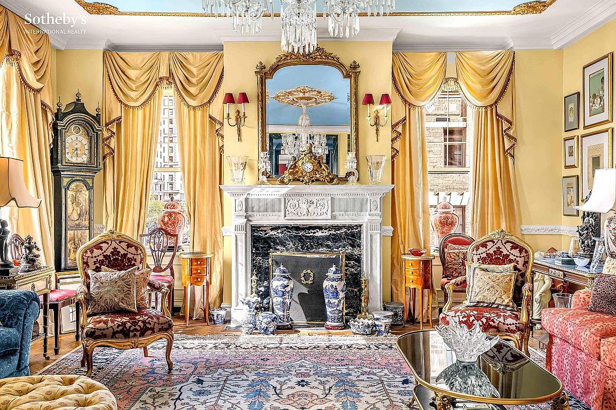 This is an opulent living room featuring a marble fireplace, ornate mirror, and luxurious gold-toned drapes. The room is furnished with antique-style chairs, a patterned rug, and various decorative items, creating a sophisticated and elegant atmosphere. The perspective is a wide shot, capturing the grandeur of the space.