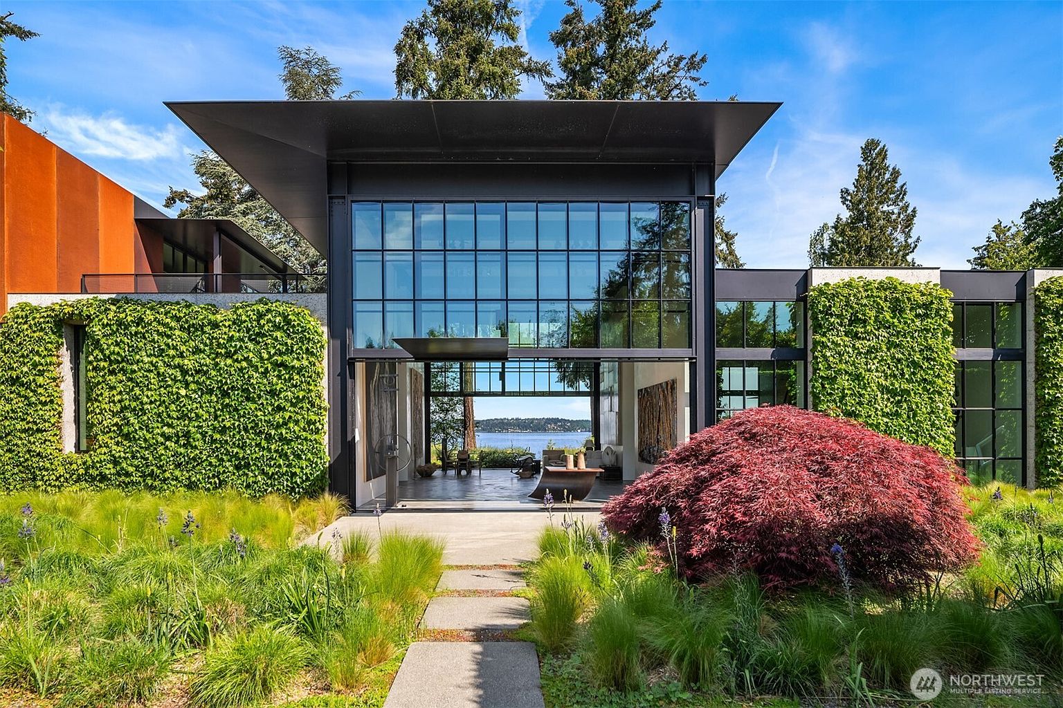This is a striking front view of a modern home, characterized by its expansive glass facade and sleek, dark roofline. Lush greenery, including climbing vines and ornamental grasses, softens the architectural lines and adds a touch of nature. A stone pathway leads to the open entryway, offering a glimpse of the interior and the water view beyond.