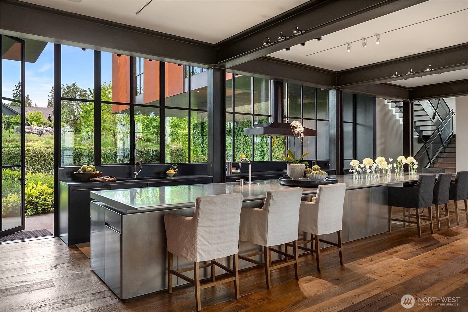 This is a modern kitchen with a large island featuring bar seating and a stainless steel countertop. The kitchen has floor-to-ceiling windows that offer a view of the outdoors, and the space is illuminated by track lighting. The flooring is hardwood, and there is a staircase visible in the background.