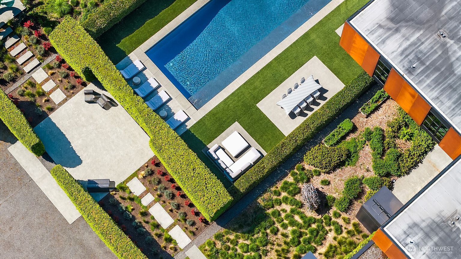 This aerial shot showcases a meticulously designed backyard with a rectangular swimming pool, manicured hedges, and various outdoor living spaces. A dining area with a long table and chairs sits adjacent to the pool, while a lounge area with a sectional sofa offers a more relaxed setting. The landscape is a mix of green lawns, stone pathways, and decorative plantings, creating a luxurious and private outdoor retreat.