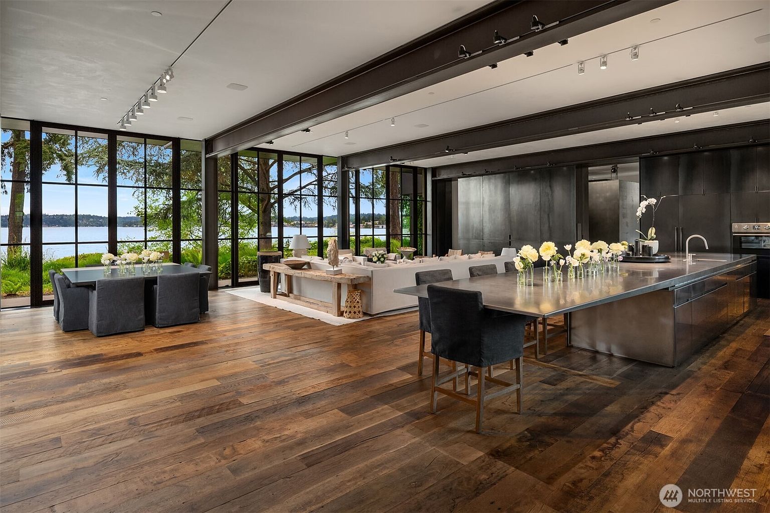 This is a spacious and modern living area with floor-to-ceiling windows offering a view of the water. The room features hardwood floors, a large sofa, a dining table, and a kitchen island with bar seating. The dark metal beams and accents add an industrial touch to the contemporary design.