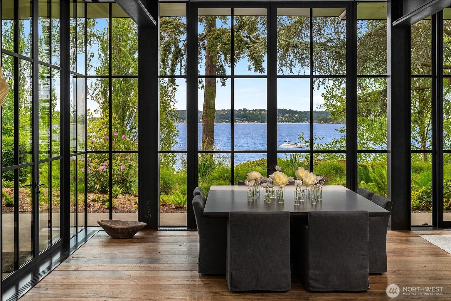 This interior shot showcases a modern dining room with a large, dark-colored dining table surrounded by upholstered chairs. The room features expansive windows with black frames, offering a scenic view of a lake and lush greenery. The hardwood flooring adds warmth to the space, creating an inviting atmosphere for dining and entertaining.