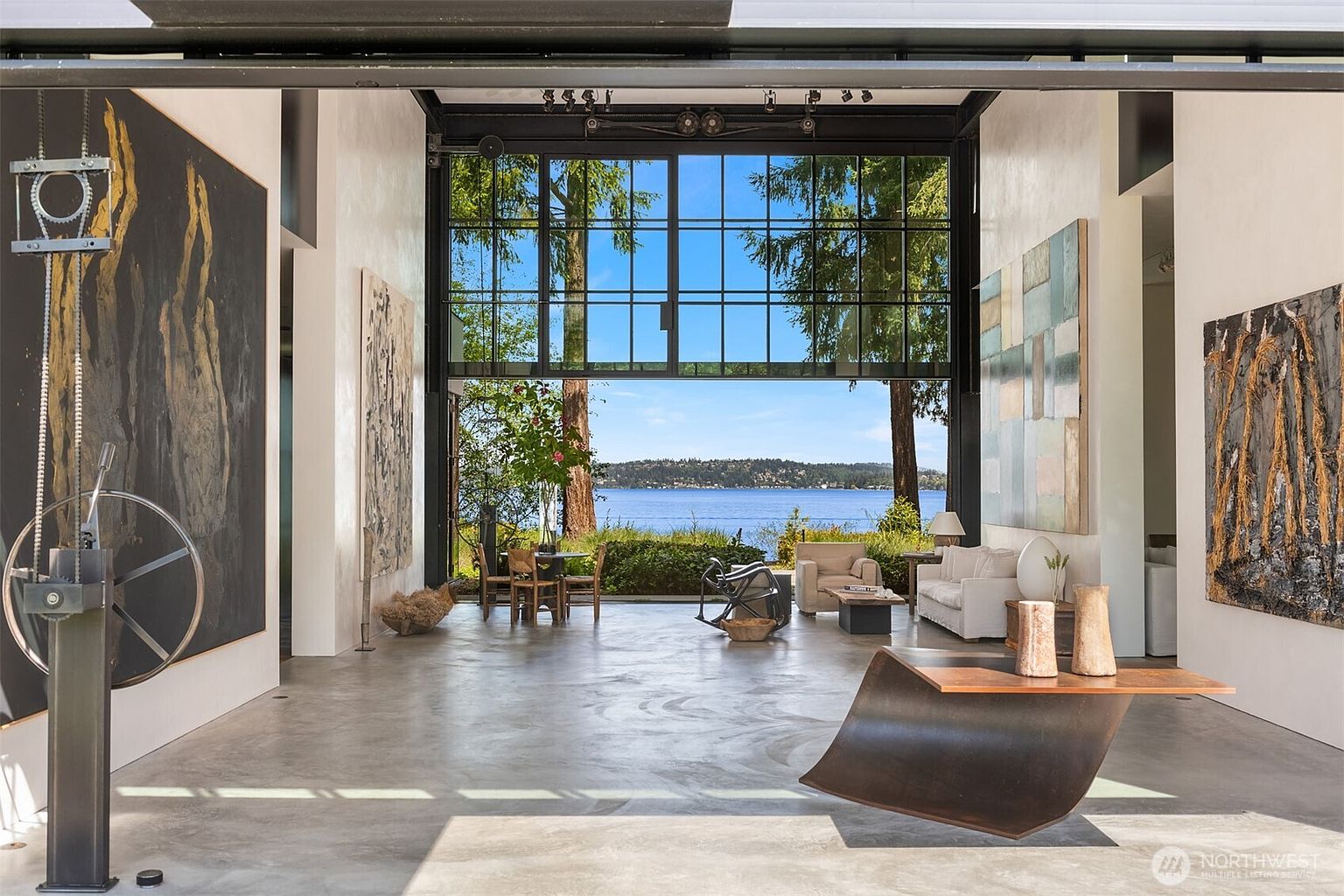 This is a spacious and modern living room with an open floor plan and a wall of windows offering a view of the water. The room features contemporary art, unique furniture pieces, and a polished concrete floor. The overall impression is one of luxury and sophistication.