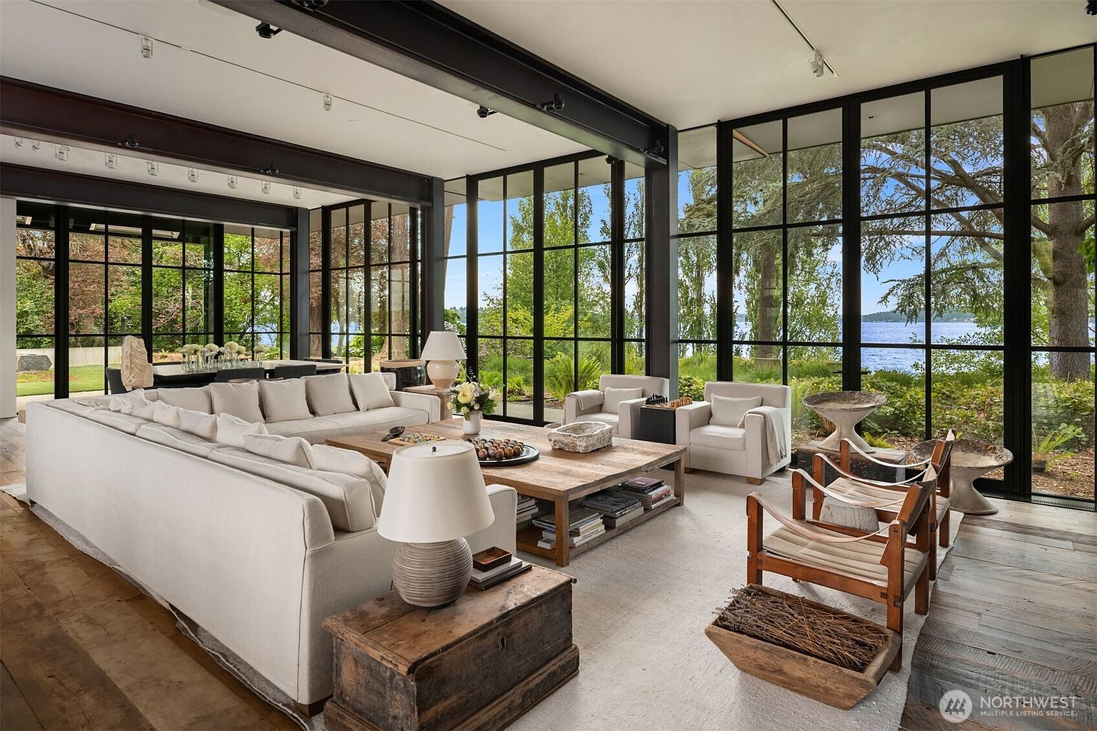 This is a spacious and luxurious living room with floor-to-ceiling windows offering stunning views of the surrounding landscape. The room features a large, comfortable sectional sofa, wooden coffee table, and stylish armchairs, creating a welcoming and sophisticated atmosphere. The hardwood floors and natural light enhance the room's appeal, making it an ideal space for relaxation and entertaining.