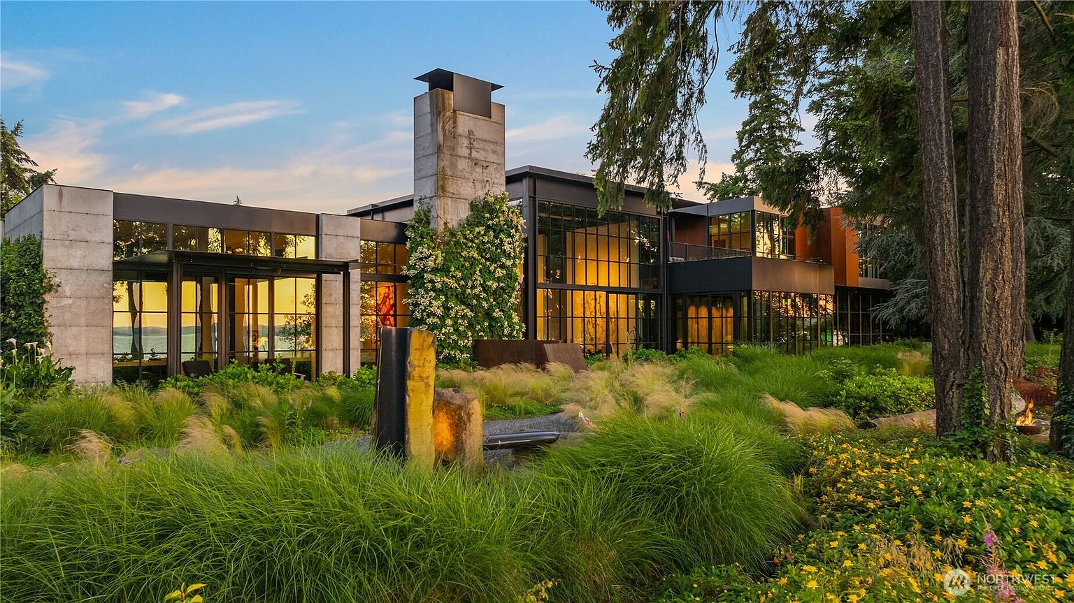 This is a striking front view of a modern home, showcasing its architectural design with a combination of concrete, glass, and metal elements. The landscaping features lush greenery and ornamental grasses, creating a serene and inviting atmosphere. Large windows reflect the warm light of the setting sun, highlighting the home's connection to its natural surroundings.