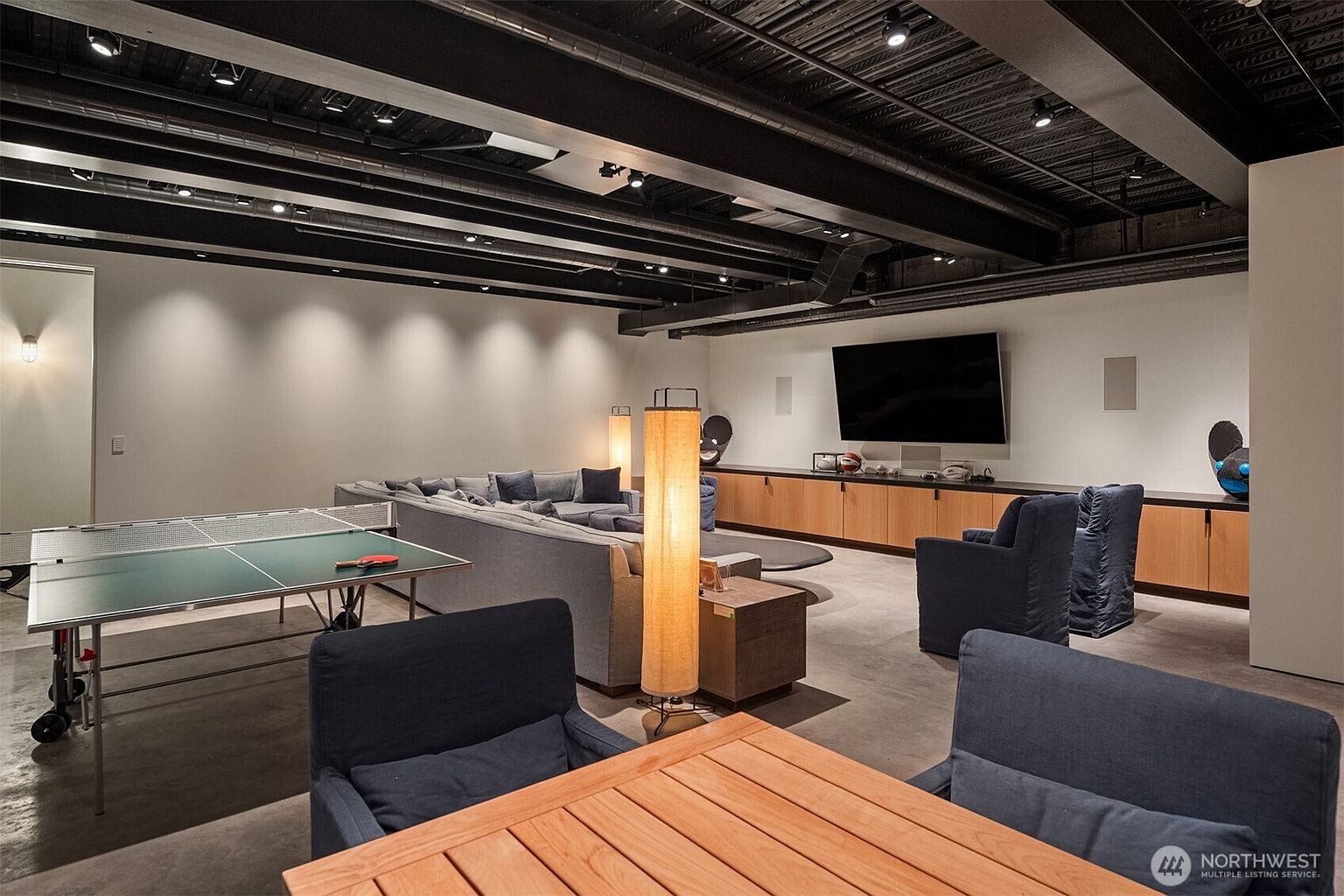 This is a spacious basement featuring a modern entertainment area with a large sectional sofa, a flat-screen TV mounted above a wooden cabinet, and stylish lighting fixtures. A ping pong table adds a recreational element to the space. The exposed ceiling beams painted in black and the concrete floor give the room a contemporary, industrial feel.
