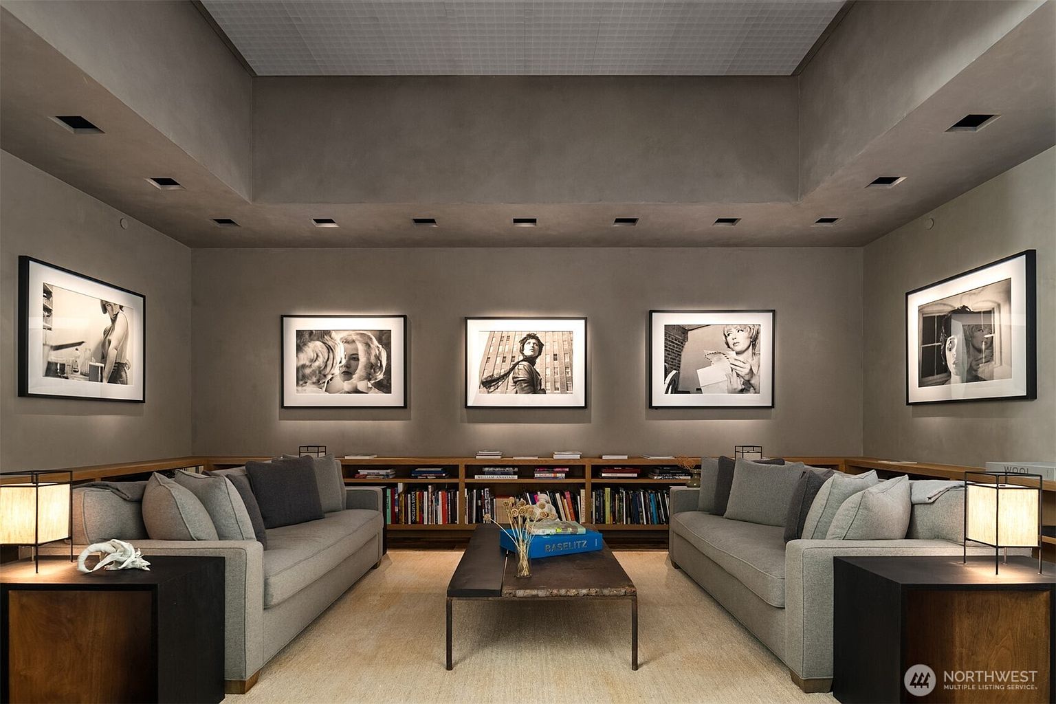 This is an interior shot of a living room featuring two gray sofas facing each other, separated by a dark wood coffee table. The room is decorated with black and white framed photographs on the walls, and a long bookshelf filled with books runs along the back wall. Two side tables with lamps sit on either side of the sofas, creating a cozy and sophisticated atmosphere.