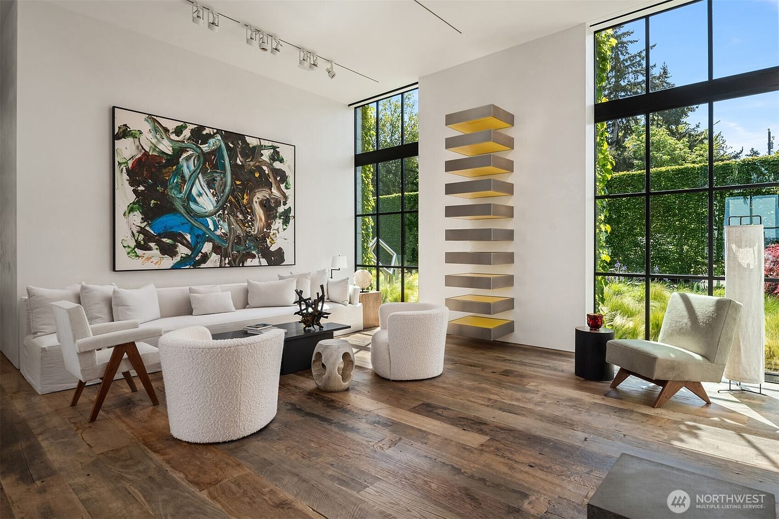 This is a bright and airy living room featuring hardwood floors, a large abstract painting, and floor-to-ceiling windows that offer a view of lush greenery. The room is furnished with a white sofa, two round white chairs, and a black coffee table, creating a modern and inviting space. A unique shelving unit adds an artistic touch to the room.