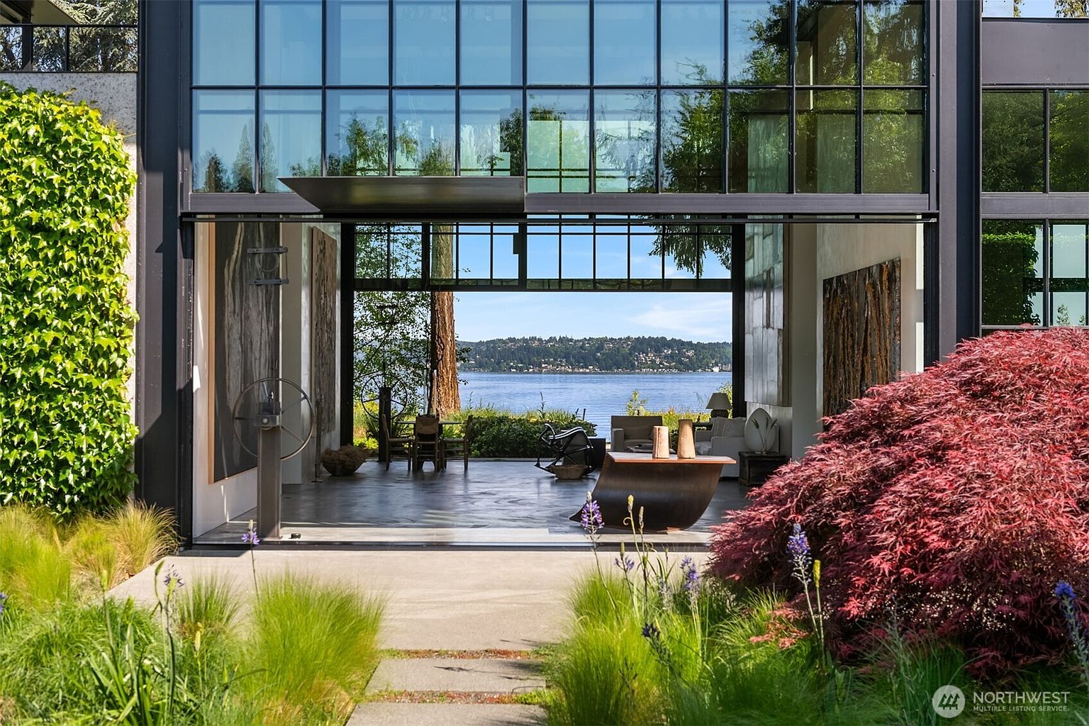 This image showcases a stunning modern home with an open-concept design that seamlessly blends indoor and outdoor living. The expansive glass walls offer breathtaking views of the water, while the lush garden and vibrant red Japanese maple create a serene and inviting atmosphere. The concrete patio extends from the interior, providing an ideal space for relaxation and entertainment.