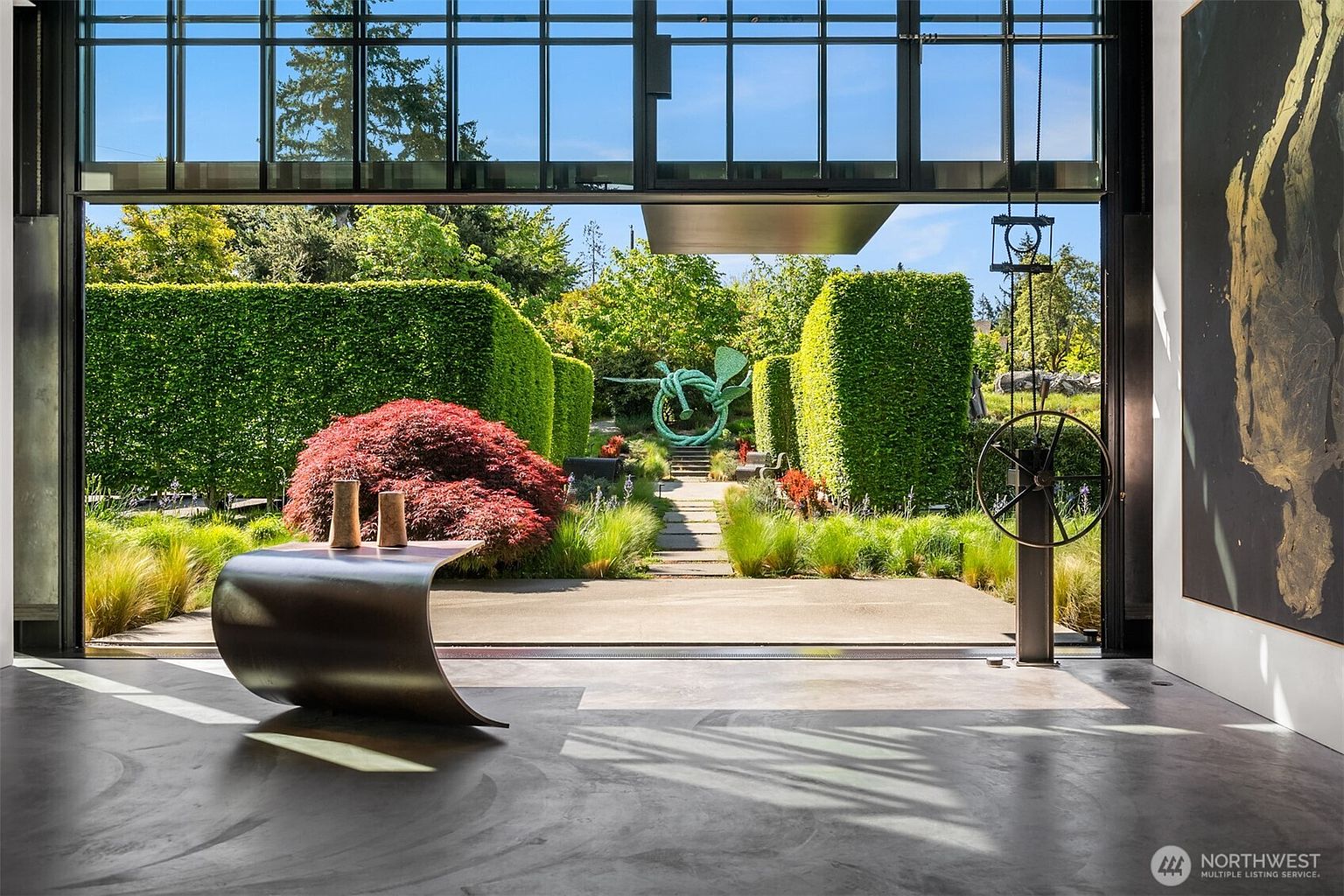 This image showcases a modern living room with an open view to a meticulously landscaped garden. The interior features a polished concrete floor and a unique, curved table, while the exterior boasts manicured hedges, a red Japanese maple, and a striking green sculpture. The large glass doors create a seamless transition between indoor and outdoor spaces, emphasizing the property's connection to nature.