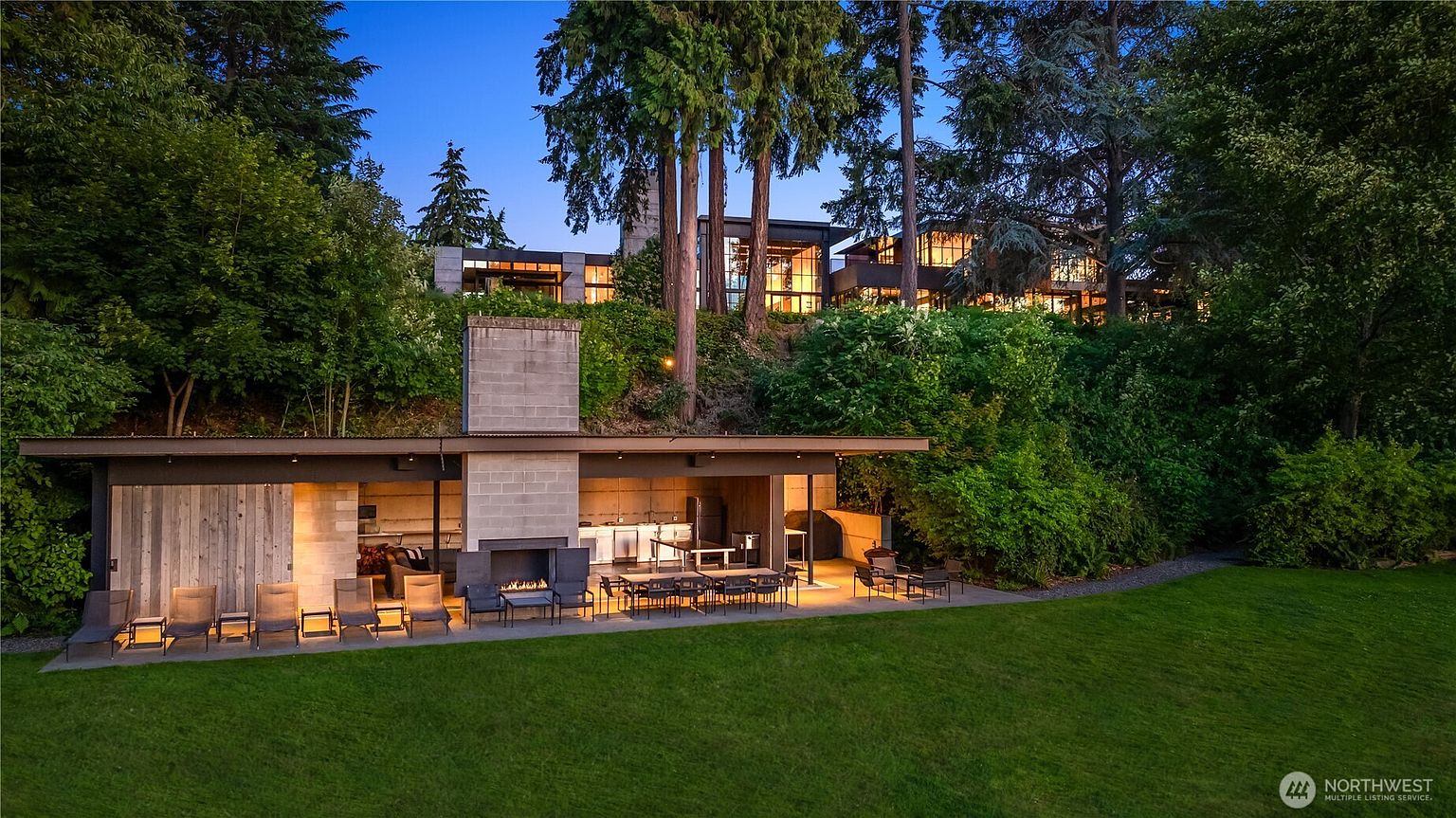 This image showcases a beautifully landscaped backyard featuring a modern outdoor kitchen and dining area. The space includes a fireplace, seating, and a dining table, all set against a backdrop of lush greenery and mature trees. The architecture of the main house is visible in the background, suggesting a luxurious and private estate.