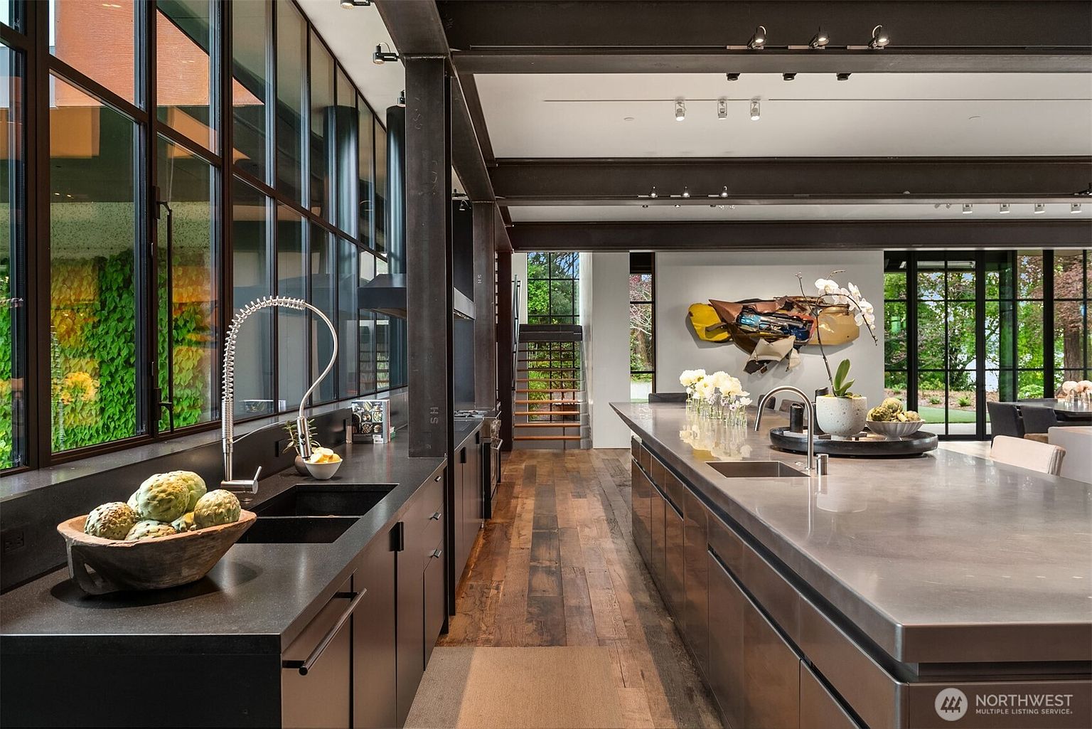 This is a modern kitchen with dark cabinetry and a large island with a stainless steel countertop. The kitchen features large windows that offer a view of the outdoors, and the hardwood floors add warmth to the space. The overall style is sleek and contemporary, with a focus on clean lines and natural light.