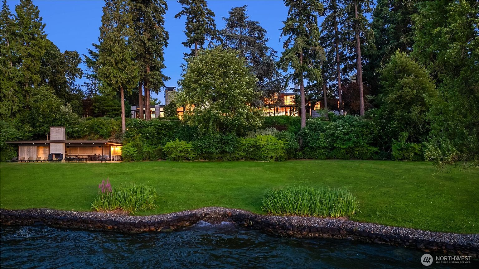 This image showcases a beautifully landscaped yard extending from a waterfront property. The lush green lawn slopes gently towards a stone-lined shoreline, with mature trees and manicured shrubs providing privacy and a sense of tranquility. A modern outdoor structure with a fireplace is visible on the left, suggesting an ideal space for entertaining.