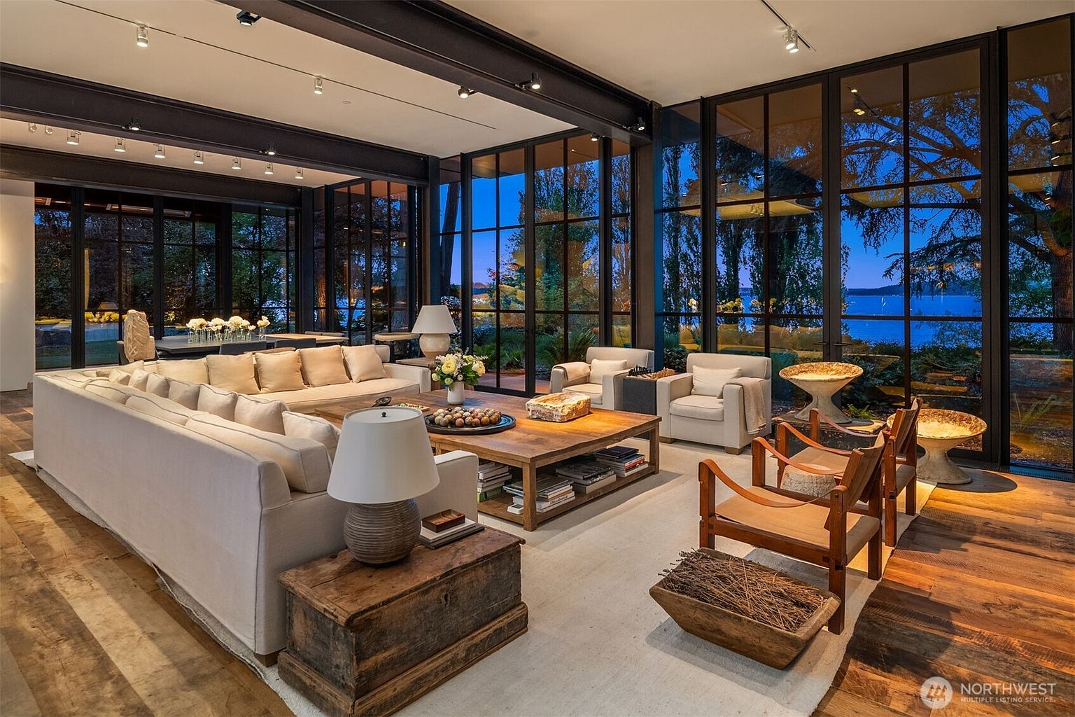 This is a spacious living room featuring a large sectional sofa, wooden coffee table, and two armchairs. The room has floor-to-ceiling windows offering a view of the outdoors. The overall style is modern and luxurious, with a focus on natural light and comfortable seating.