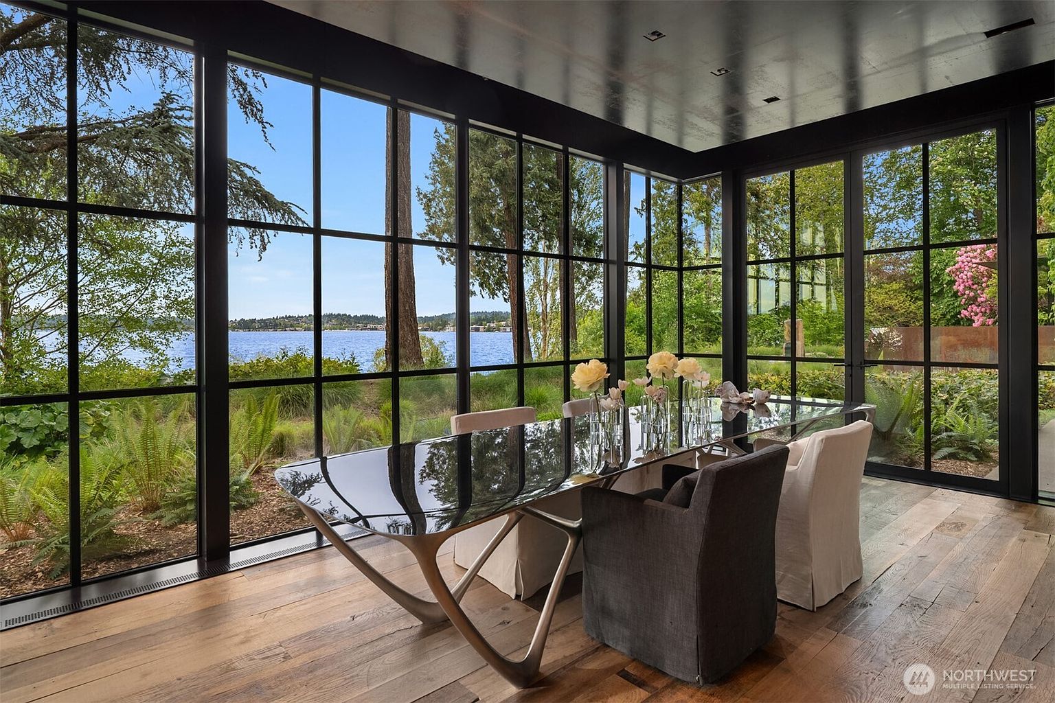 This is an interior shot of a dining room featuring a modern glass-top table with unique metal legs, surrounded by upholstered chairs. Large windows with black frames offer a stunning view of a lake and lush greenery, creating a bright and airy atmosphere. The hardwood flooring adds warmth to the space, complementing the contemporary design.