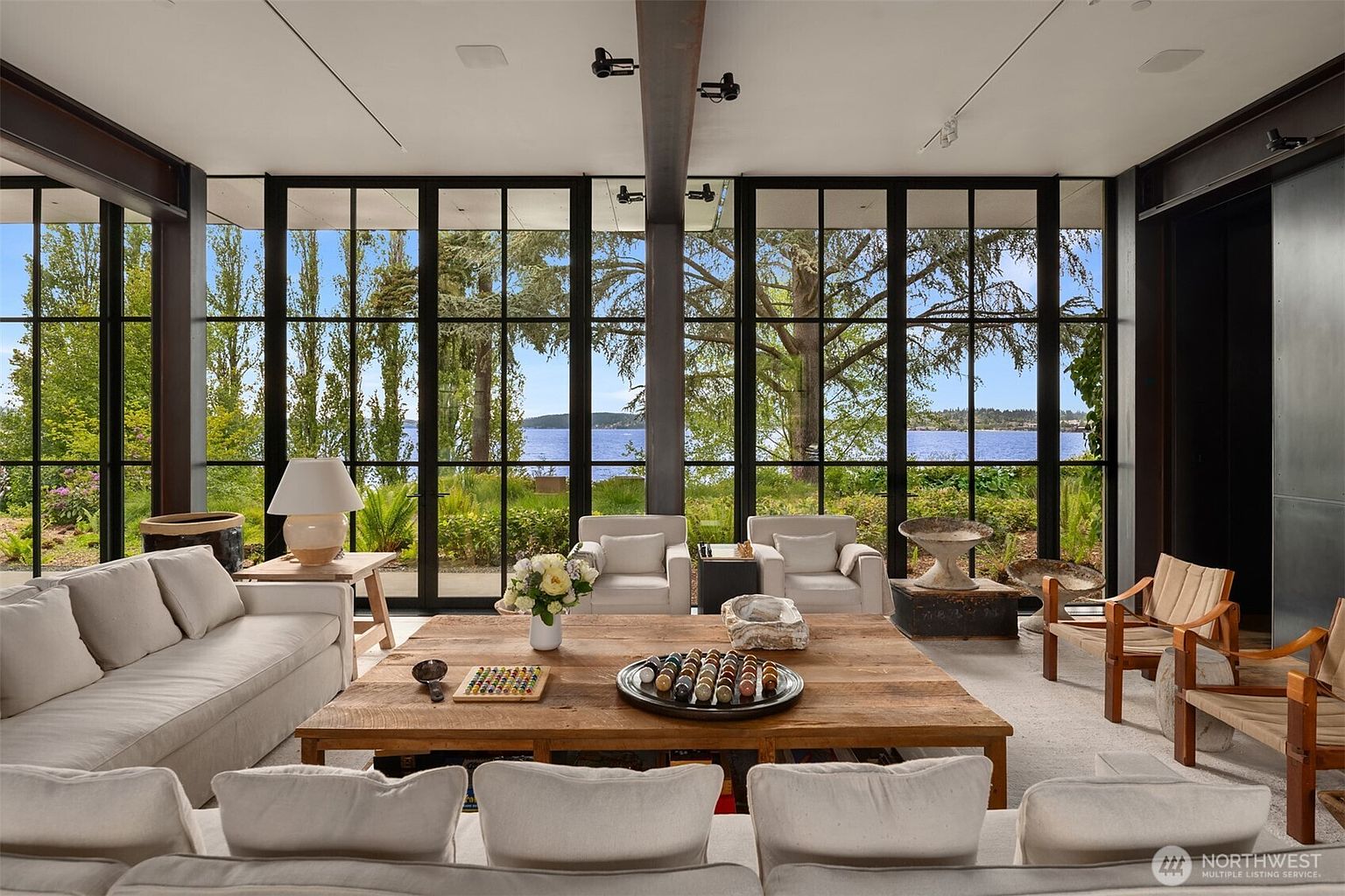 This is a bright and airy living room with large windows offering a view of the water and lush greenery. The room features neutral-toned sofas, wooden chairs, and a large wooden coffee table, creating a comfortable and inviting space. The design emphasizes natural light and a connection to the outdoors, perfect for relaxation and entertaining.