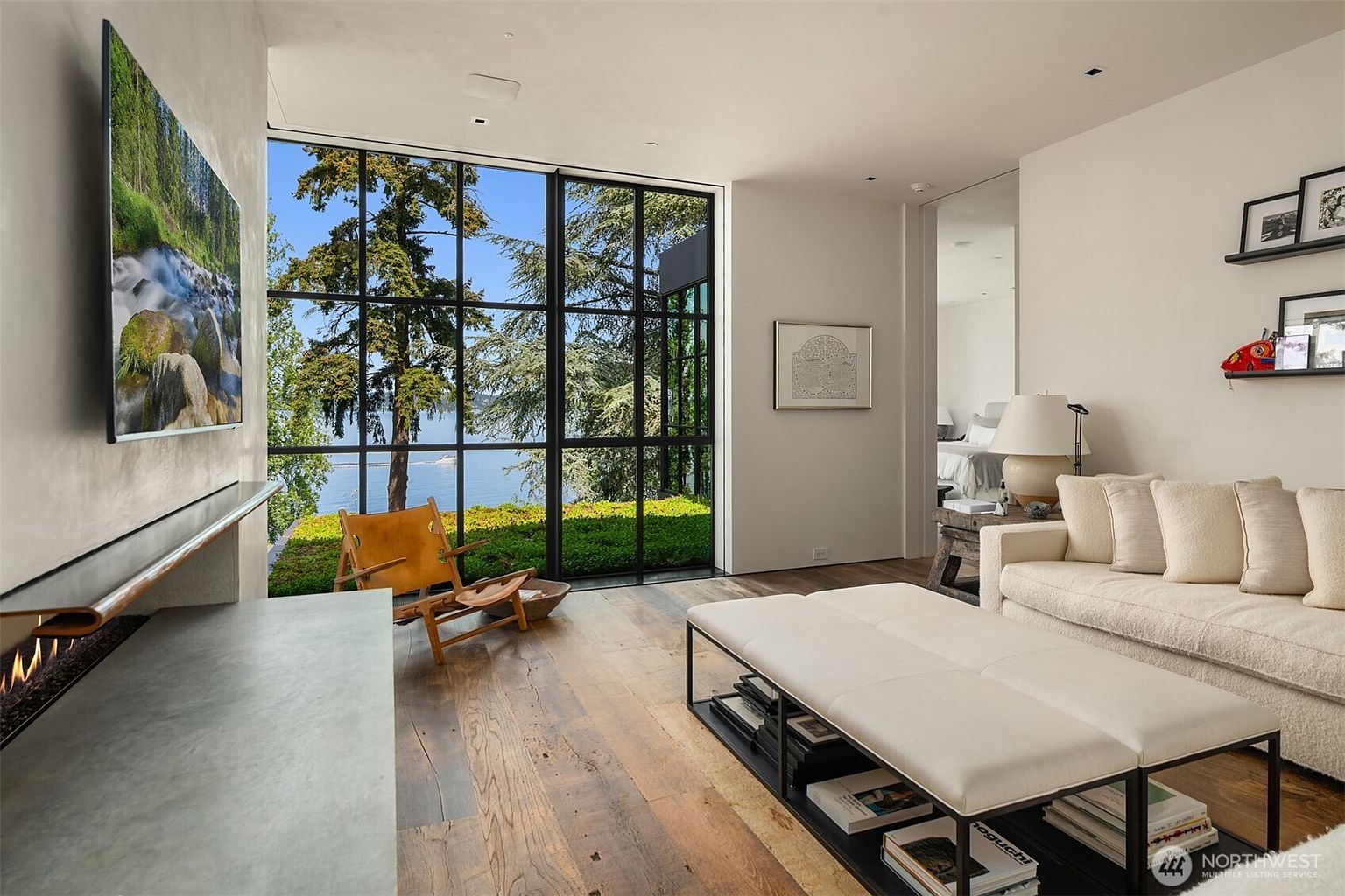 This is an interior shot of a modern living room featuring a large window with a view of the water and trees. The room is furnished with a light-colored sofa, a bench with books underneath, and a unique wooden chair. A fireplace and a television are mounted on the wall, adding to the room's contemporary design.
