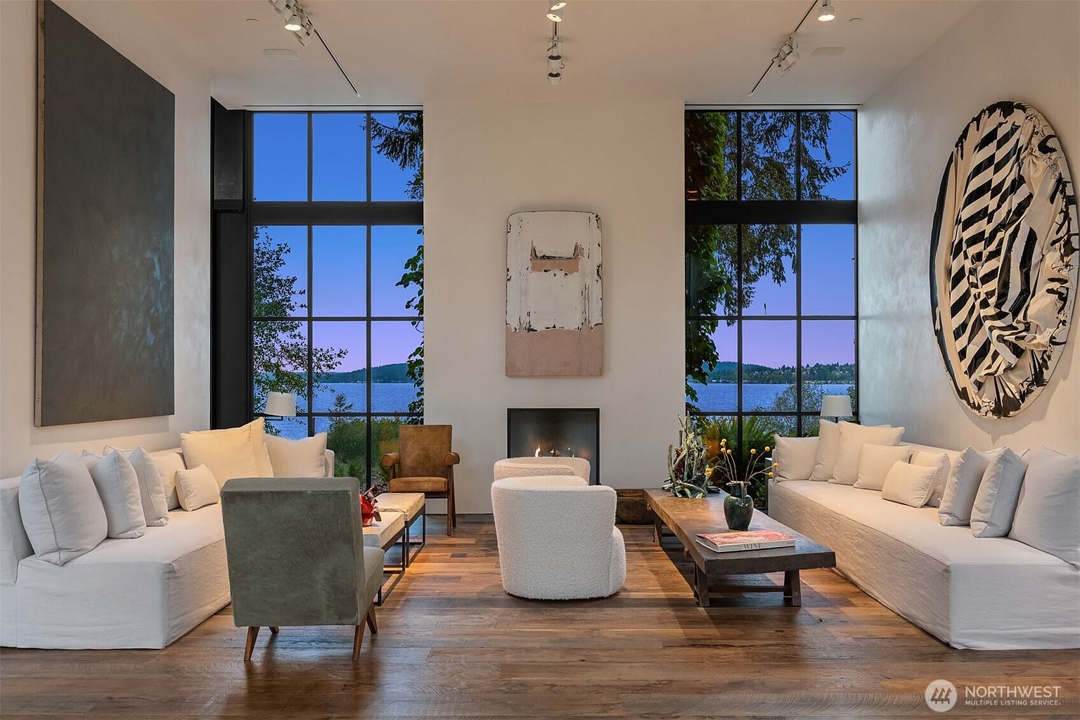 This is a spacious living room featuring hardwood floors, large windows with lake views, and modern art pieces. The room is furnished with comfortable white sofas, a textured armchair, and a rustic coffee table, creating a cozy yet sophisticated atmosphere. A fireplace adds warmth and a focal point to the room.