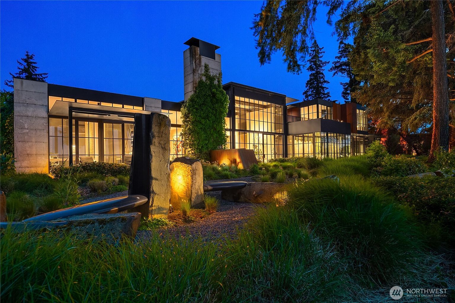 This is a captivating front view of a modern home at dusk, showcasing its architectural design with large windows and a prominent chimney. The landscape features a tranquil water feature with stone accents and lush greenery, creating an inviting and serene atmosphere. The warm interior lighting contrasts beautifully with the cool blue of the evening sky.