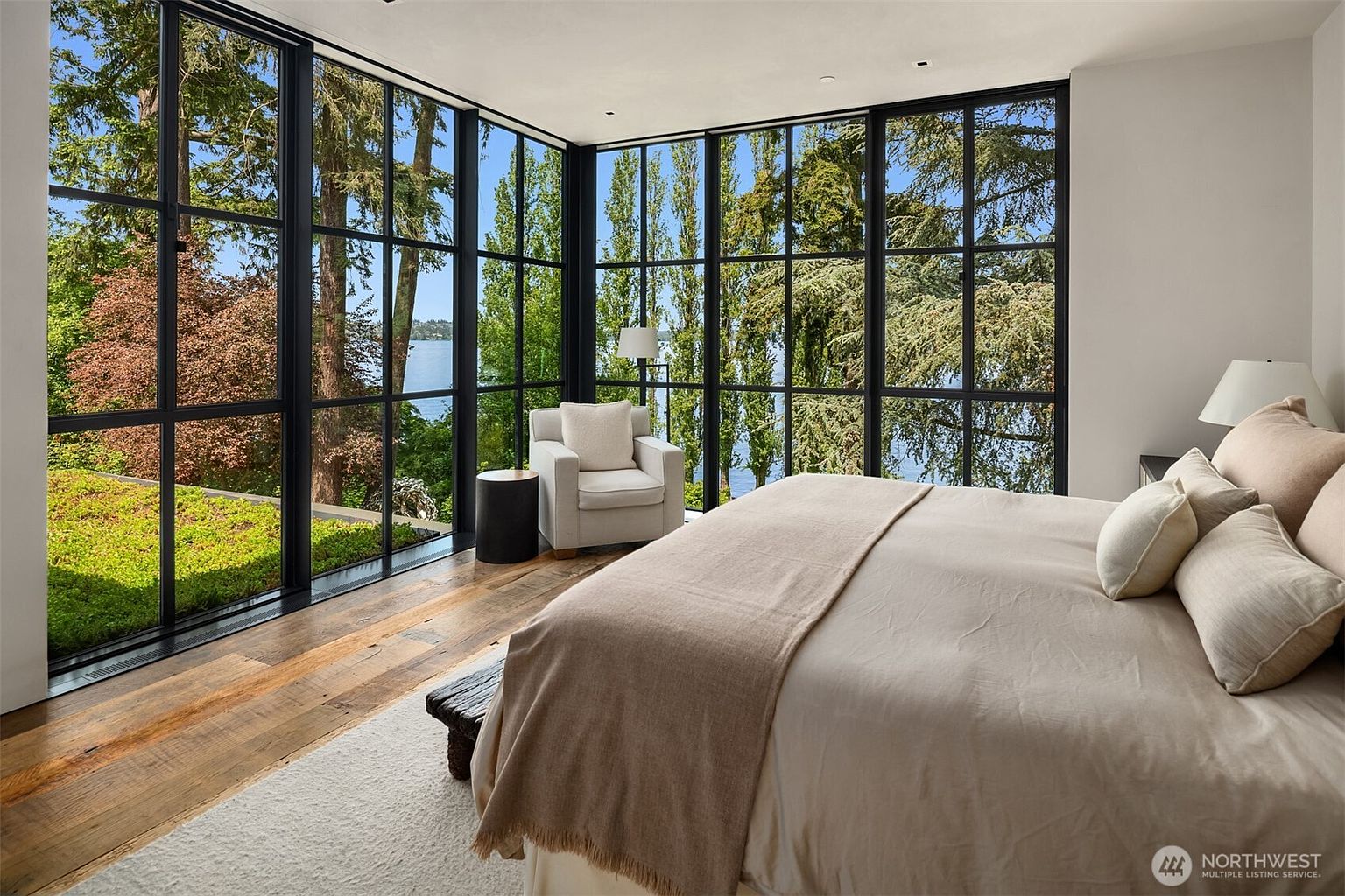 This is a bright and airy primary bedroom featuring floor-to-ceiling windows with black frames that offer stunning views of the surrounding greenery and water. The room is furnished with a large bed covered in neutral linens and pillows, a side table, and a comfortable armchair, creating a serene and inviting atmosphere. The hardwood floors and soft rug add warmth and texture to the space.