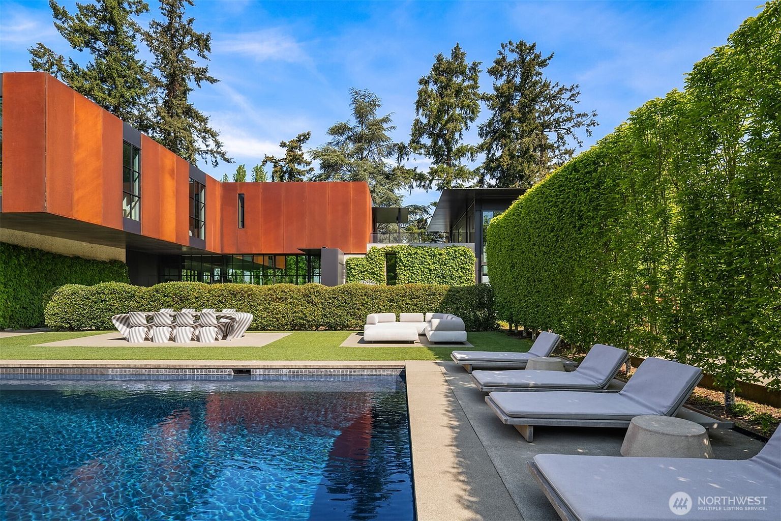 This image showcases a luxurious backyard with a modern architectural home as a backdrop. A pristine pool takes center stage, complemented by stylish lounge chairs and a comfortable outdoor seating area. The well-manicured lawn and lush greenery create a serene and private oasis, perfect for relaxation and entertainment.