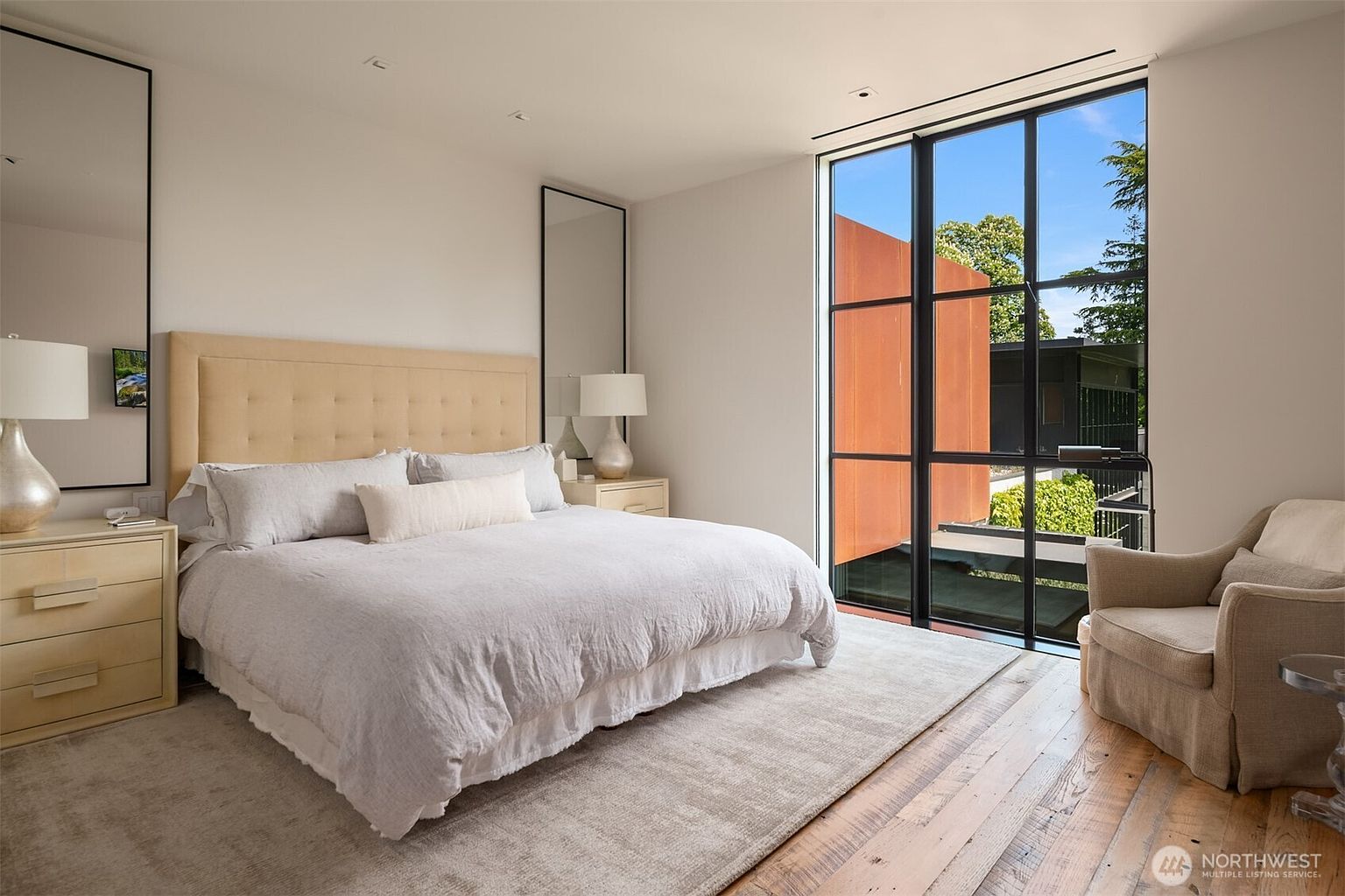 This is a bright and airy primary bedroom featuring a large bed with a neutral-toned headboard and light-colored bedding. The room has a modern aesthetic with clean lines, large windows offering natural light and views, and minimalist decor including two tall mirrors and matching nightstands with lamps. The hardwood floors and area rug add warmth and texture to the space.