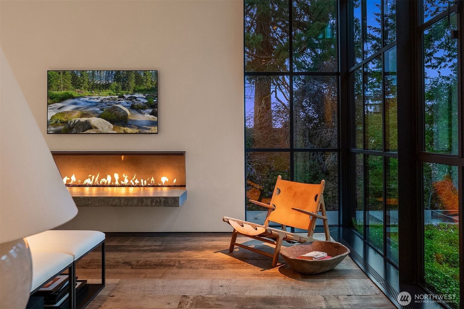 This interior shot showcases a modern living room with a minimalist aesthetic. A sleek, linear fireplace is mounted beneath a nature-themed artwork, while a stylish leather chair sits near a large window offering a view of the outdoors. The hardwood flooring and natural light create a warm and inviting atmosphere.