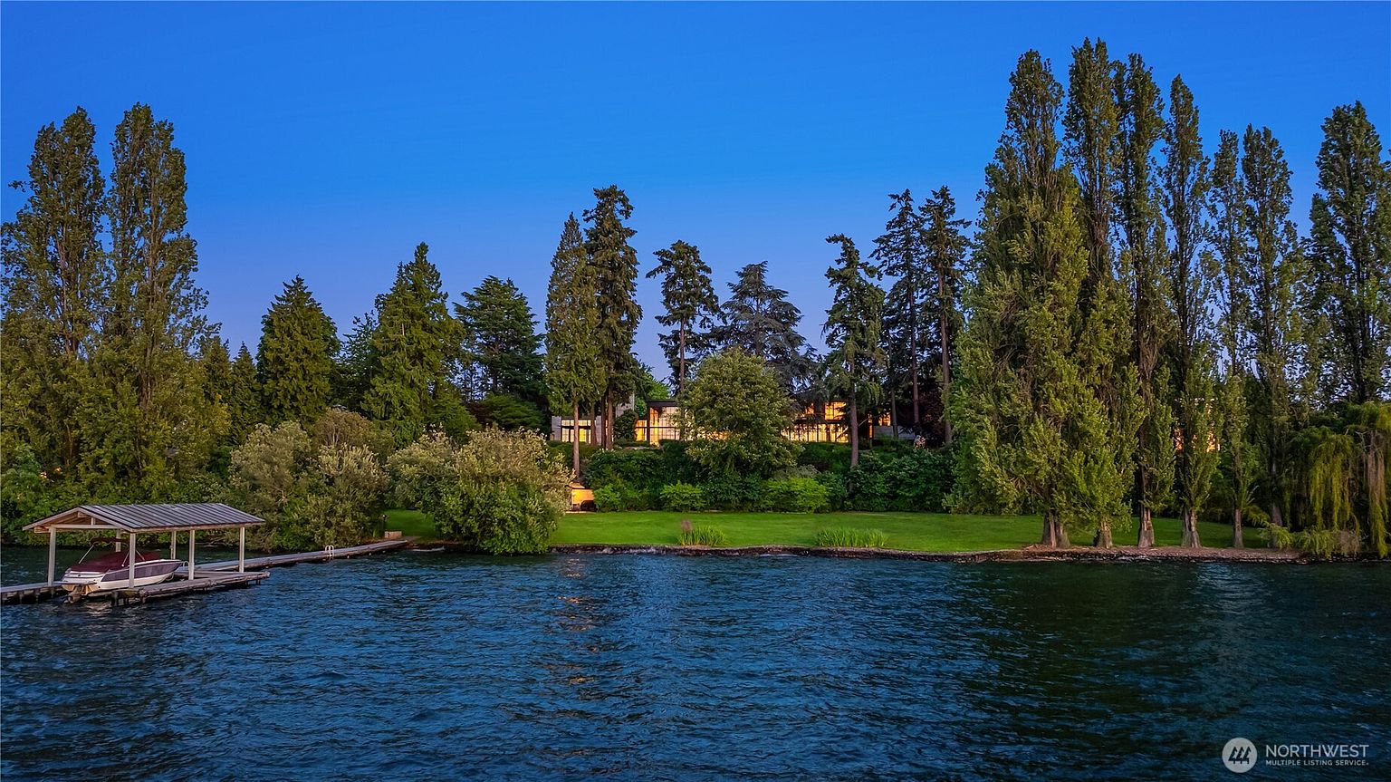 This image showcases the rear of a waterfront property, featuring a well-maintained lawn that slopes down to the water's edge. The house is nestled among mature trees, providing privacy and a serene backdrop. A private dock with a boat lift extends into the calm water, enhancing the property's appeal for boating enthusiasts.
