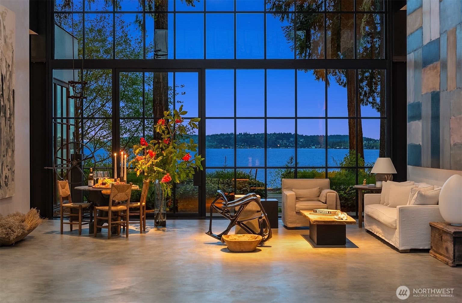 This is an interior shot of a luxurious living room with floor-to-ceiling windows offering a stunning view of a lake. The room features a dining area with a round table and chairs, a modern chair, and a comfortable seating area with a sofa and armchair. The overall style is modern and elegant, creating a serene and inviting atmosphere.