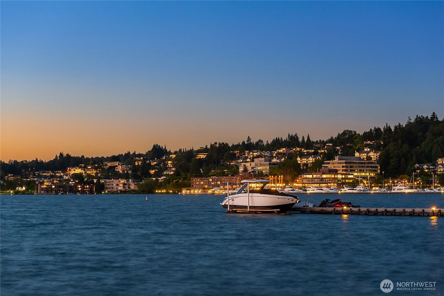 This stunning exterior shot captures a serene waterfront scene at twilight. A sleek boat is docked at a pier, with the lights of waterfront homes and buildings twinkling in the background. The calm water and clear sky create a peaceful and luxurious ambiance, perfect for showcasing a high-end property with water access.