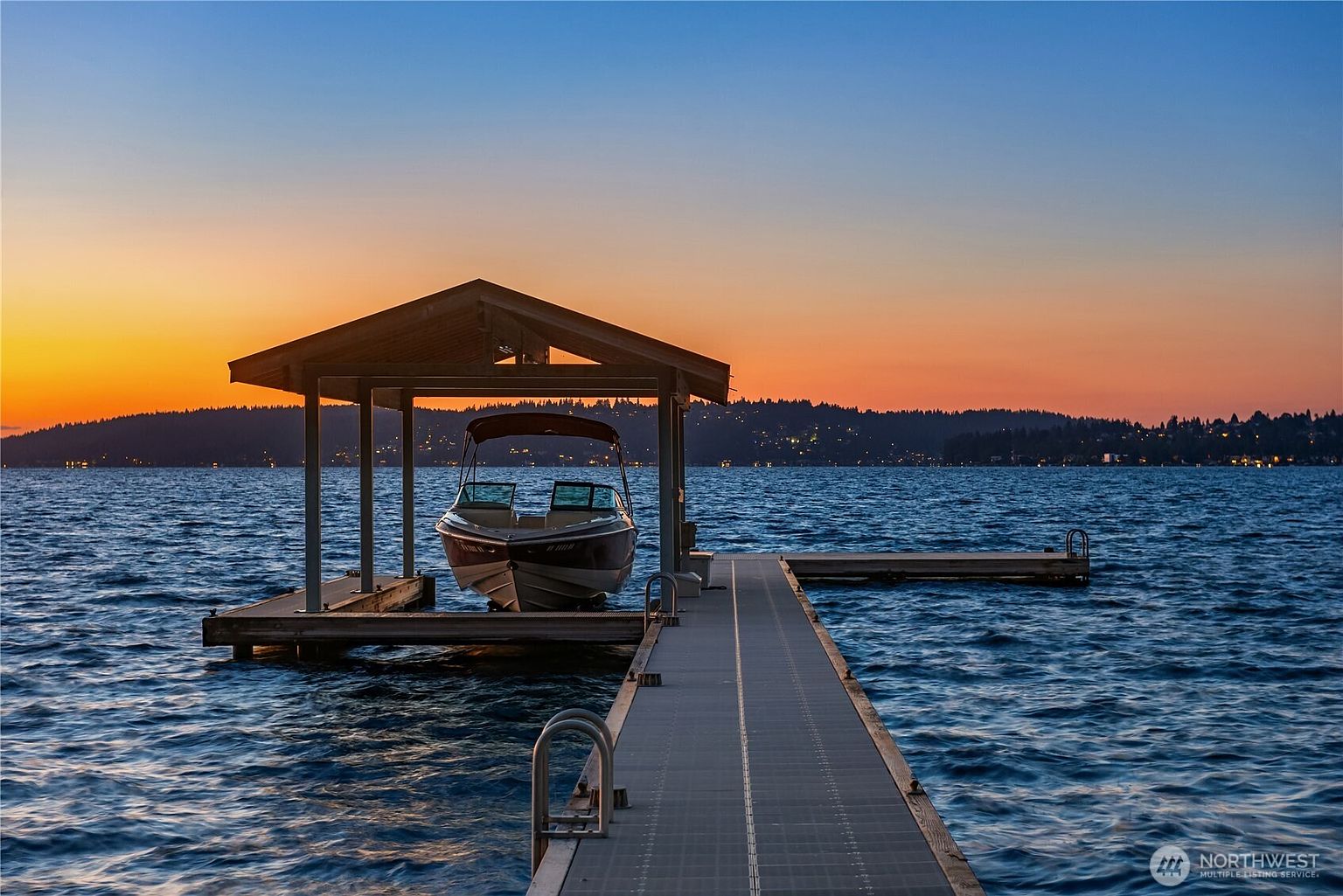 The image showcases a serene waterfront property at sunset, featuring a private dock with a boat lift and a covered boat. The dock extends into the calm water, reflecting the vibrant colors of the sky. The scene evokes a sense of tranquility and luxury, highlighting the property's access to water activities and stunning views.