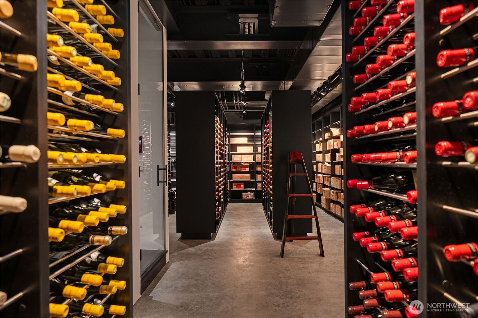 This is a well-organized wine cellar featuring rows of wine bottles with yellow and red capsules stored on metal racks. A wooden ladder stands in the center aisle, providing access to higher shelves. The cellar has a modern design with dark shelving and a concrete floor, creating a sophisticated and temperature-controlled environment for wine storage.