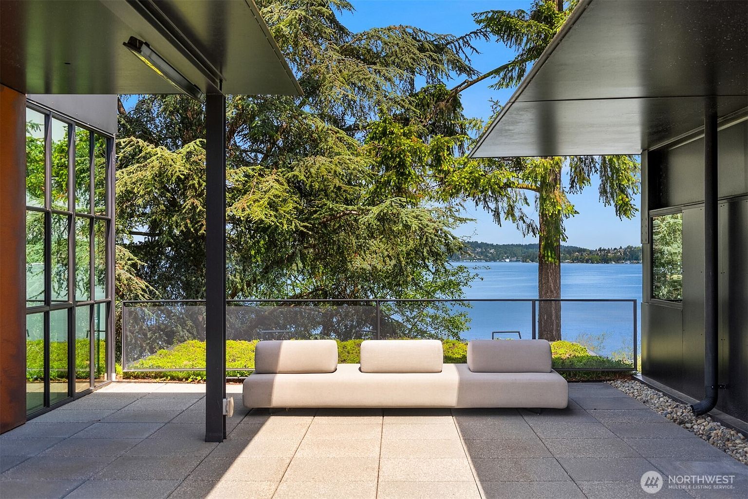 This image showcases a modern patio or deck area with a sleek, minimalist design. A long, low-profile sofa provides seating, set against a backdrop of lush greenery and a serene water view. The patio features clean lines, a neutral color palette, and a glass railing that maximizes the scenic vista, creating an inviting outdoor living space.