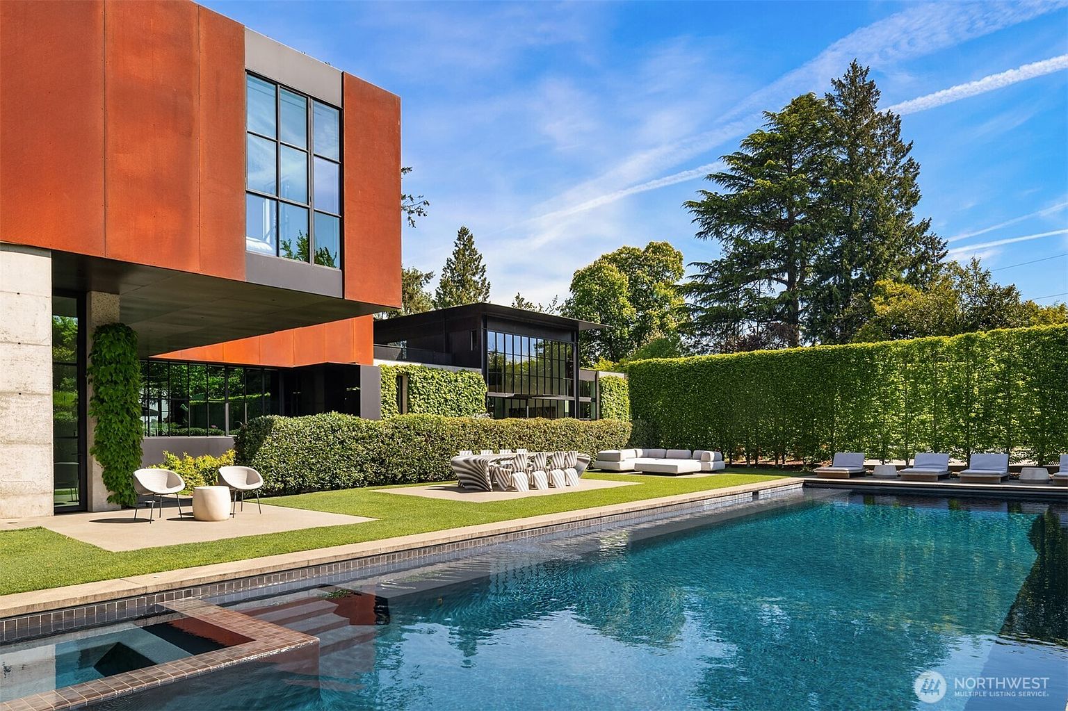 This image showcases a luxurious modern home with a stunning pool and spa area. The architecture features a combination of materials, including rust-colored panels and large windows. The pool is surrounded by manicured lawns, hedges, and stylish outdoor furniture, creating an inviting and upscale outdoor living space.