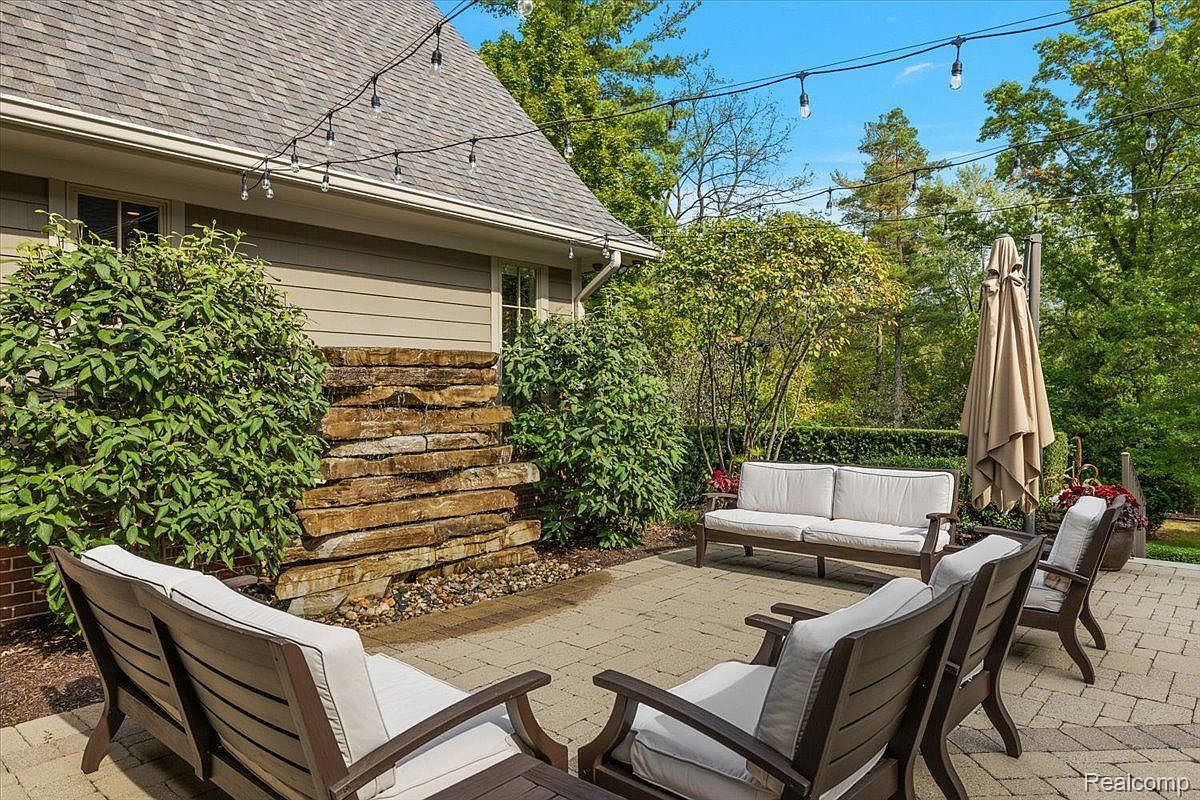 This image showcases a beautifully designed outdoor patio area, featuring comfortable seating arrangements with cushioned chairs and a sofa. A unique stone water feature adds a touch of elegance and tranquility to the space, while string lights overhead create a warm and inviting ambiance. The patio is paved with stone and surrounded by lush greenery, offering a private and relaxing outdoor retreat.